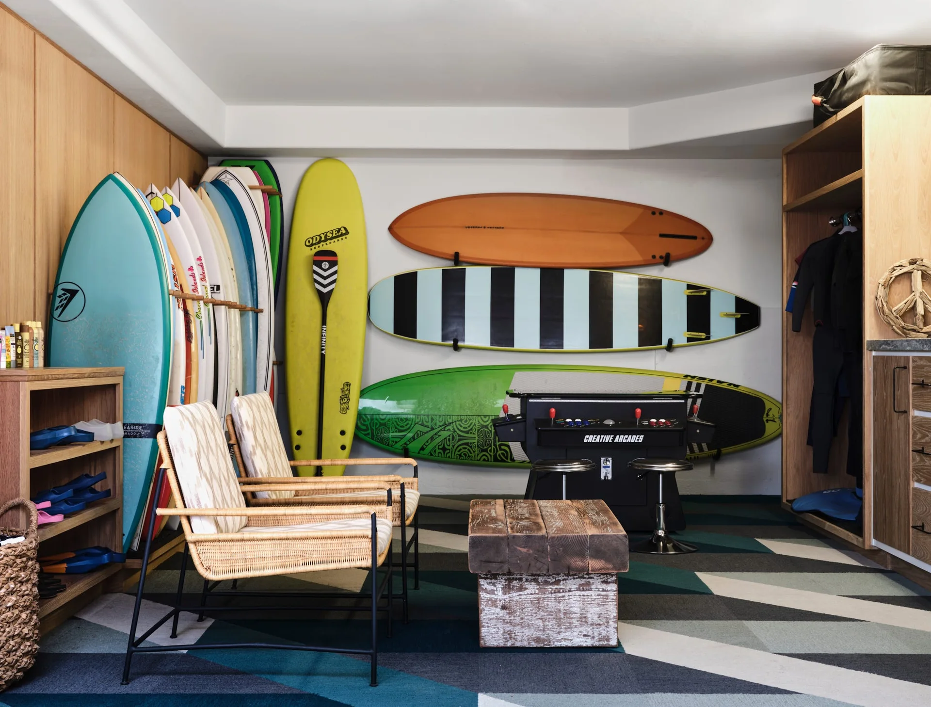 A coastal mudroom featuring colorful surfboards mounted on white walls, two rattan armchairs, a wood block coffee table, and a geometric patterned carpet.