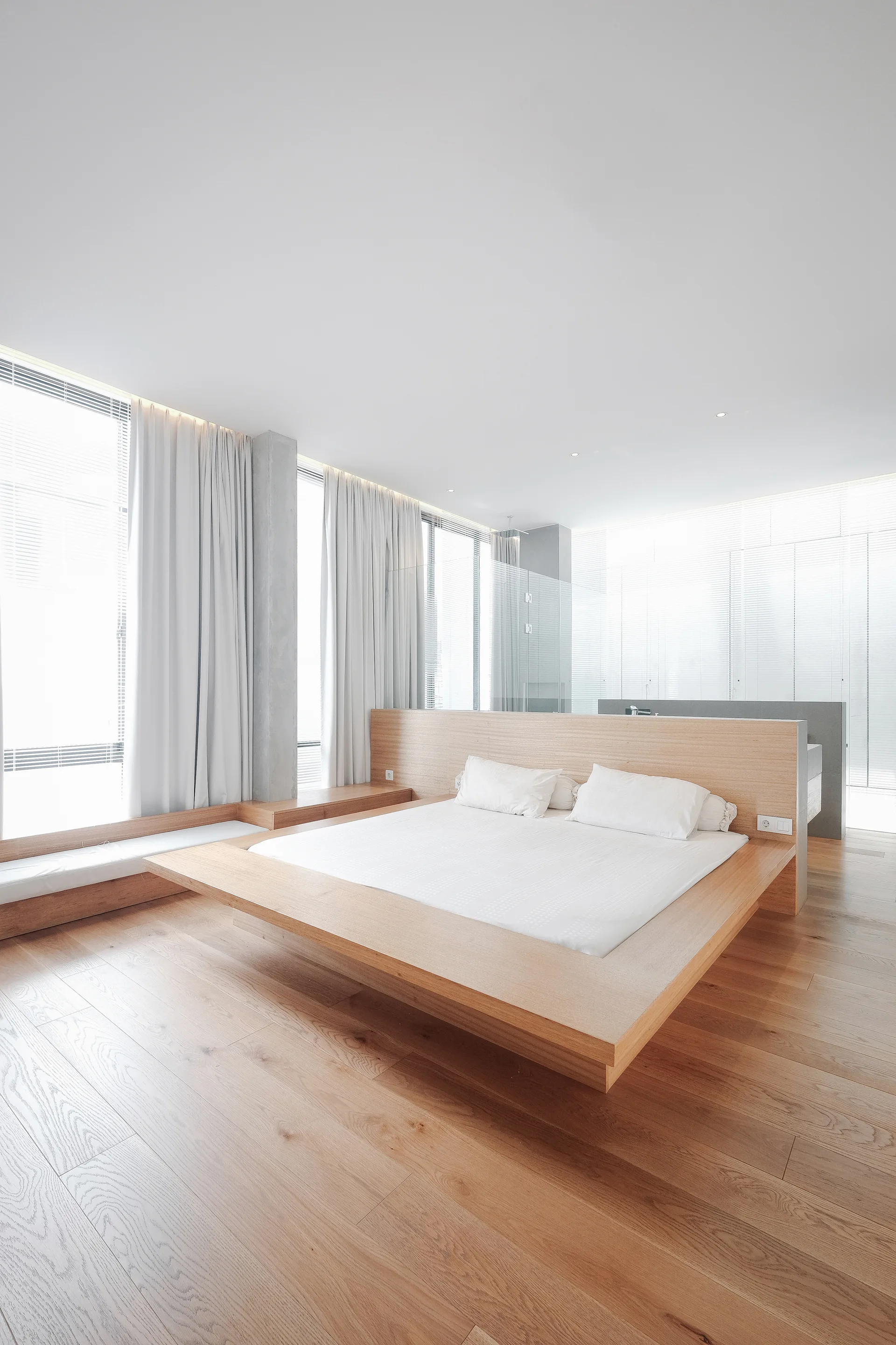 A minimalist bedroom features a low wood platform bed, a built-in wooden headboard partition, and sheer white curtains over large windows.