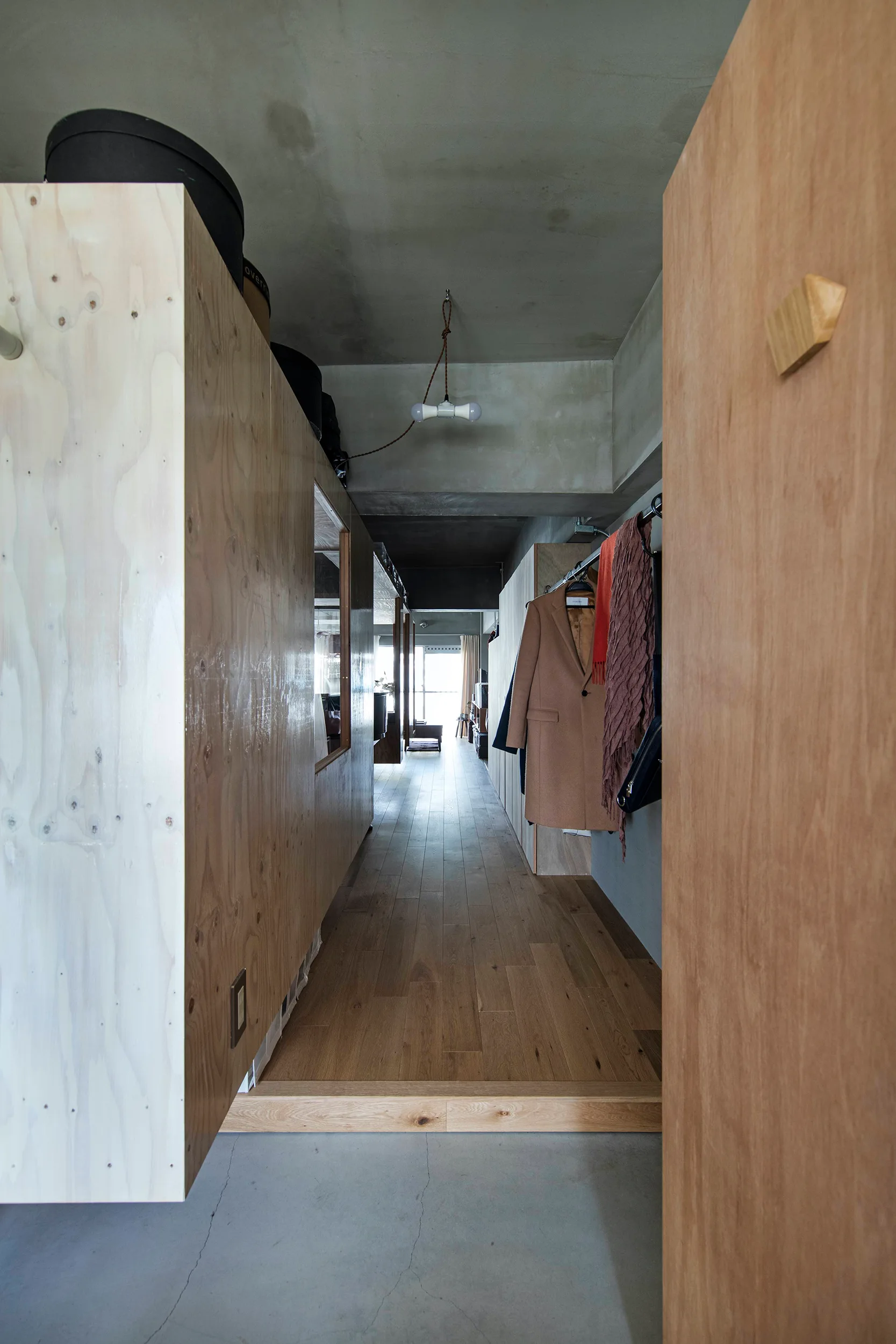 A minimalist hallway features a large plywood storage unit, wood plank floors, an exposed concrete ceiling, and a simple clothes rack on the wall.