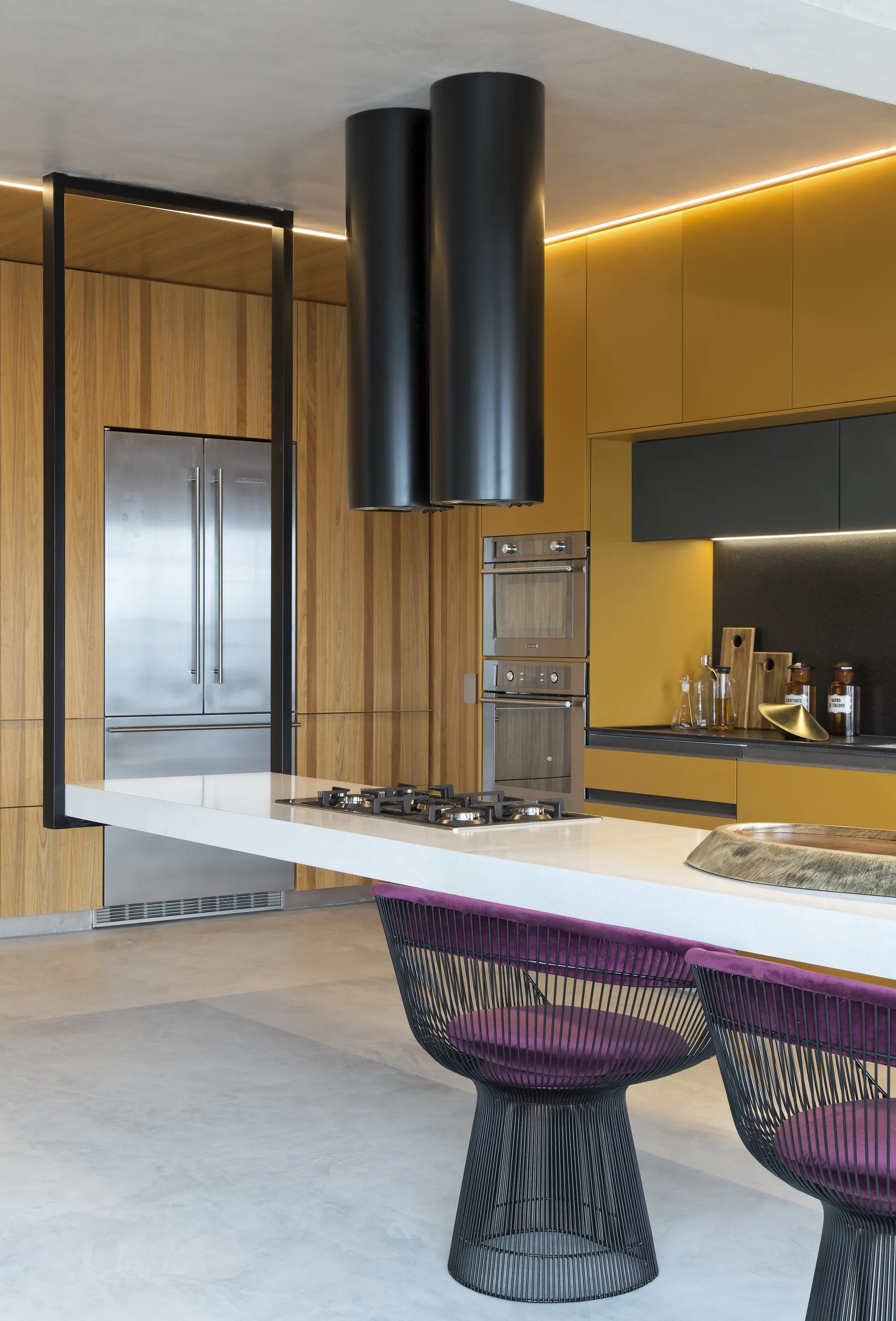 A modern kitchen featuring a cantilevered white island with purple-cushioned wire stools, mustard yellow cabinets, and two black cylindrical range hoods.
