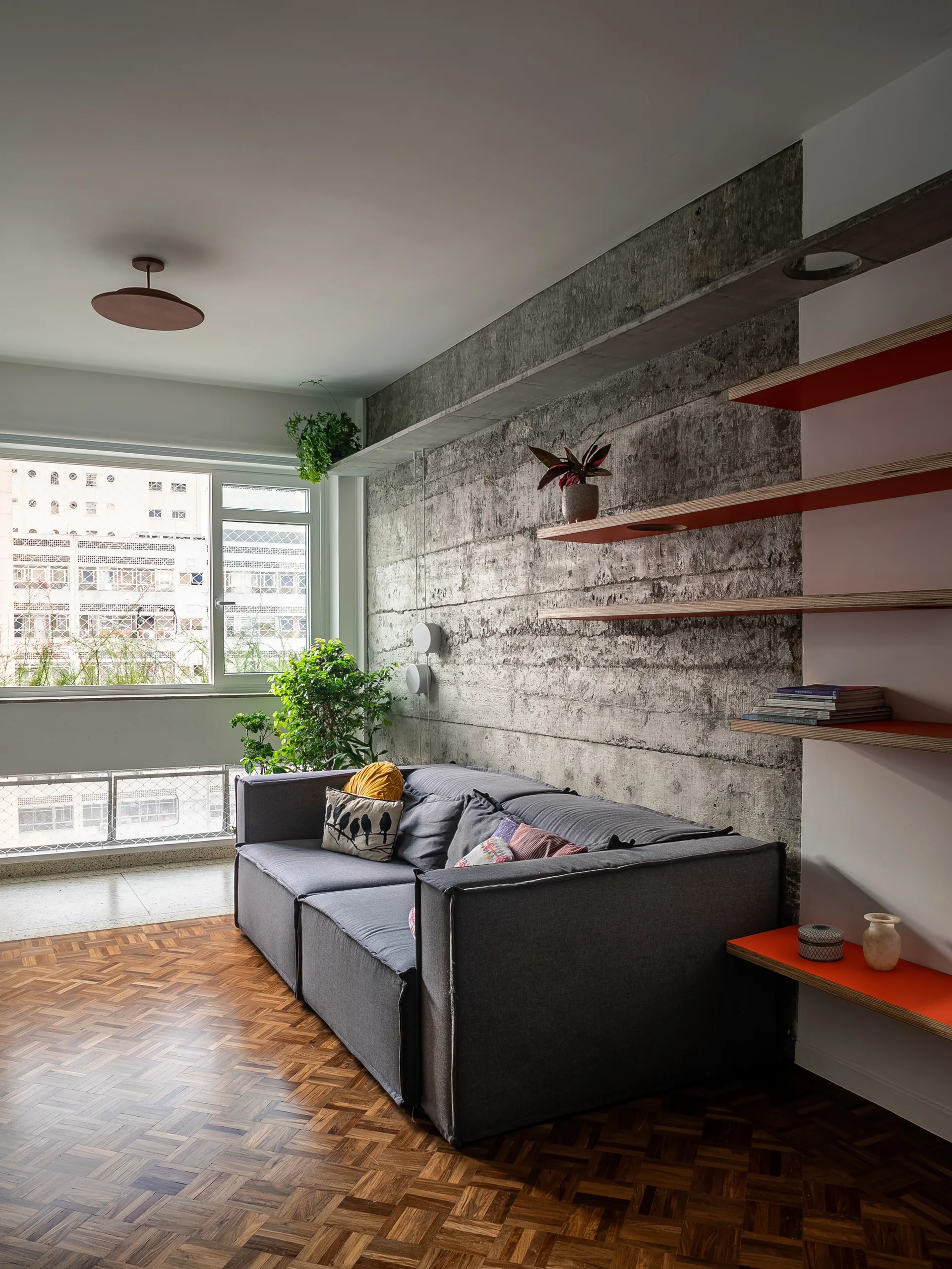 A modern grey sofa stands against a raw concrete wall fitted with orange floating shelves next to a warm wood herringbone parquet floor.