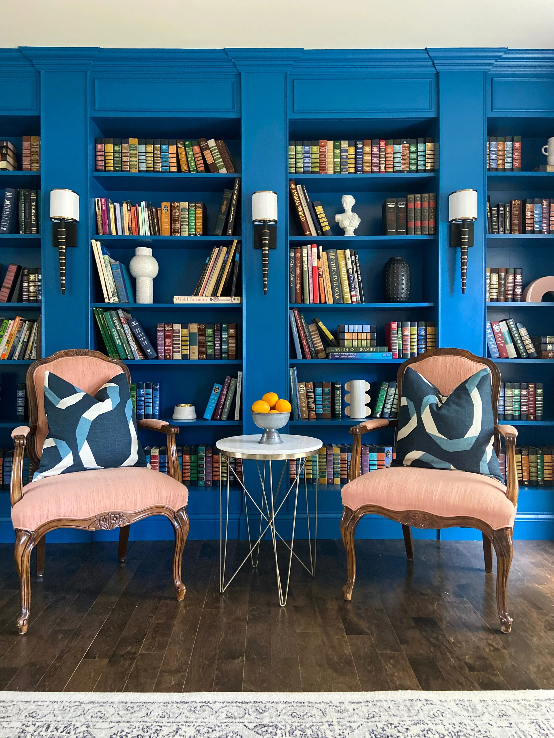 Two pink armchairs with blue geometric pillows sit in front of a wall-to-wall blue built-in bookshelf filled with books and decor.