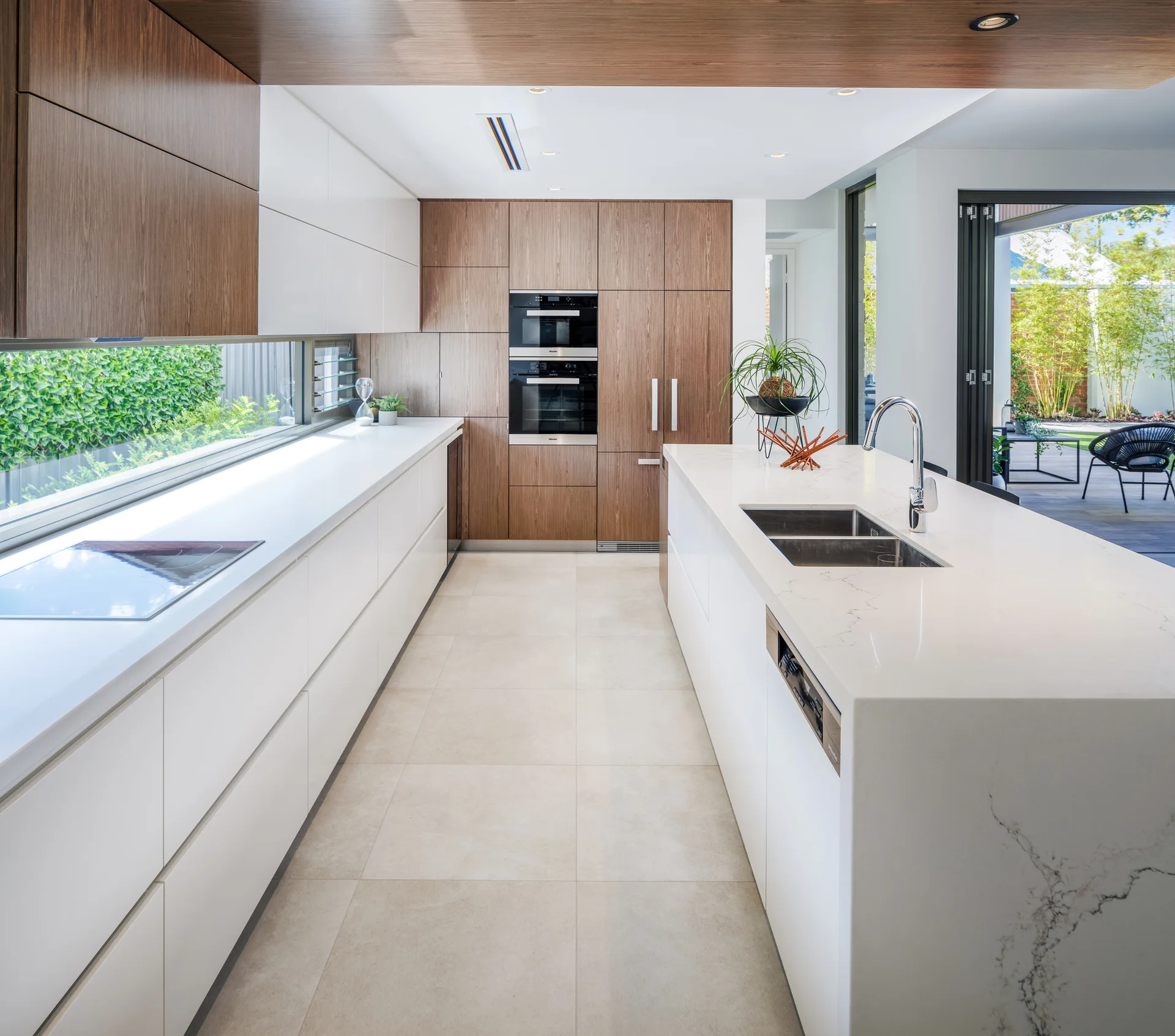 A modern kitchen with a large white stone island, wood-toned cabinets, and a long horizontal backsplash window opening to outdoor greenery.