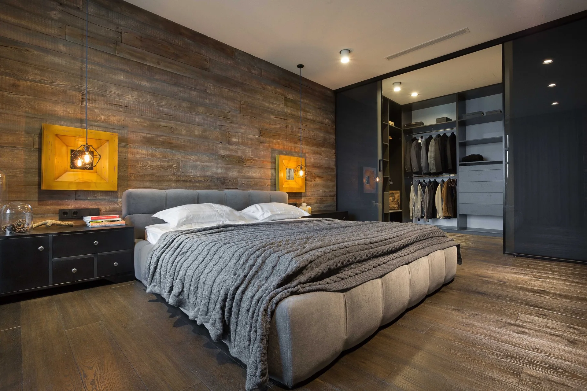 A contemporary bedroom features an upholstered grey bed, horizontal wood-plank wall cladding, black nightstands, and a walk-in wardrobe with glass doors.