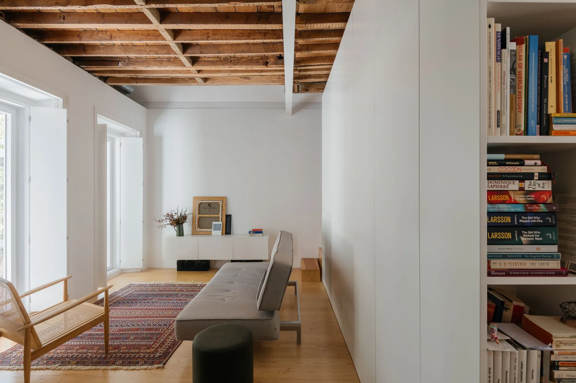 A minimalist living room features exposed wood ceiling joists, a grey sofa, a patterned rug, and a large white built-in storage wall.