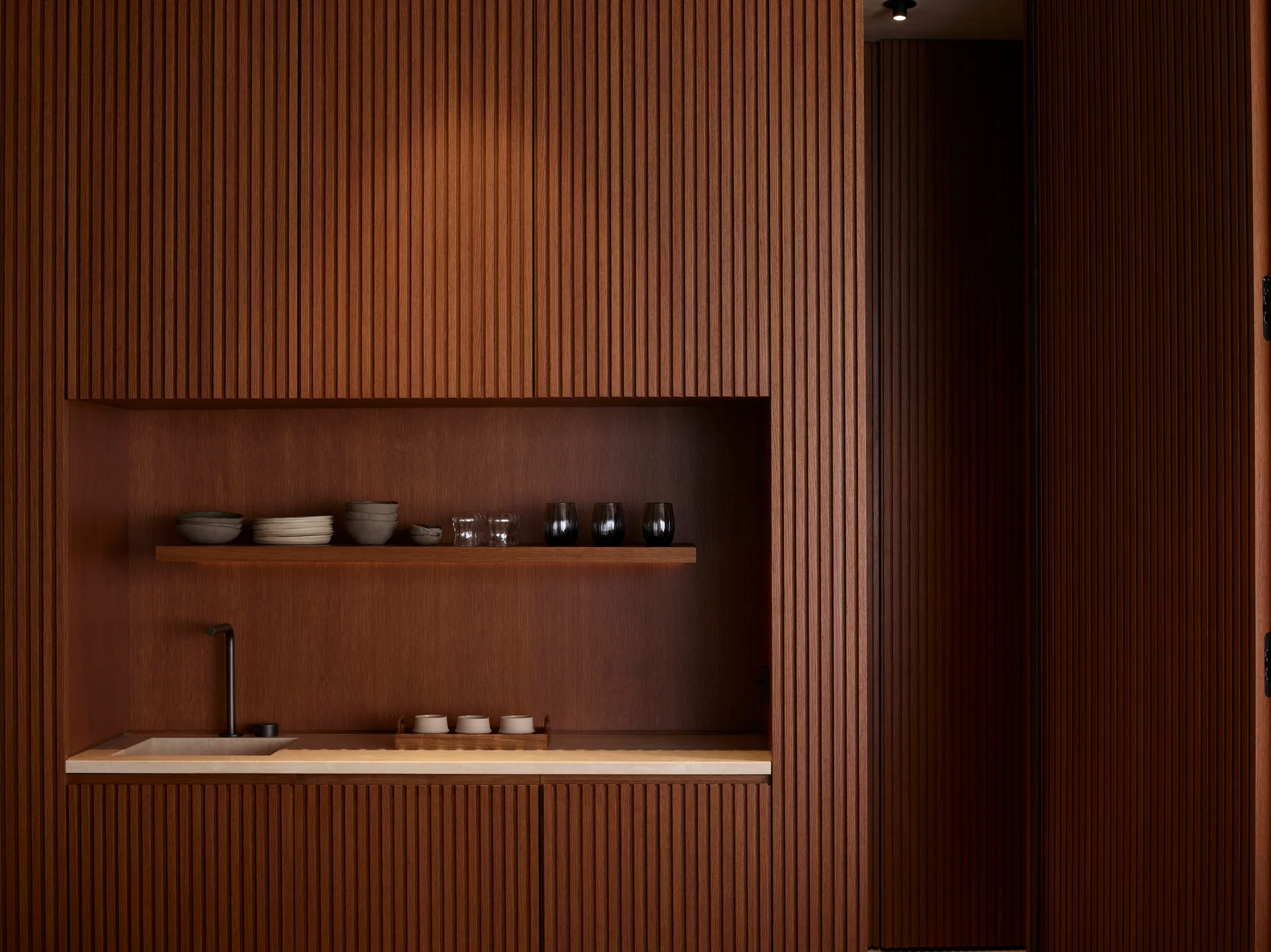 A wall of vertically ribbed wood cabinetry features a recessed nook with a stone countertop, an integrated sink with a black faucet, and a floating shelf.