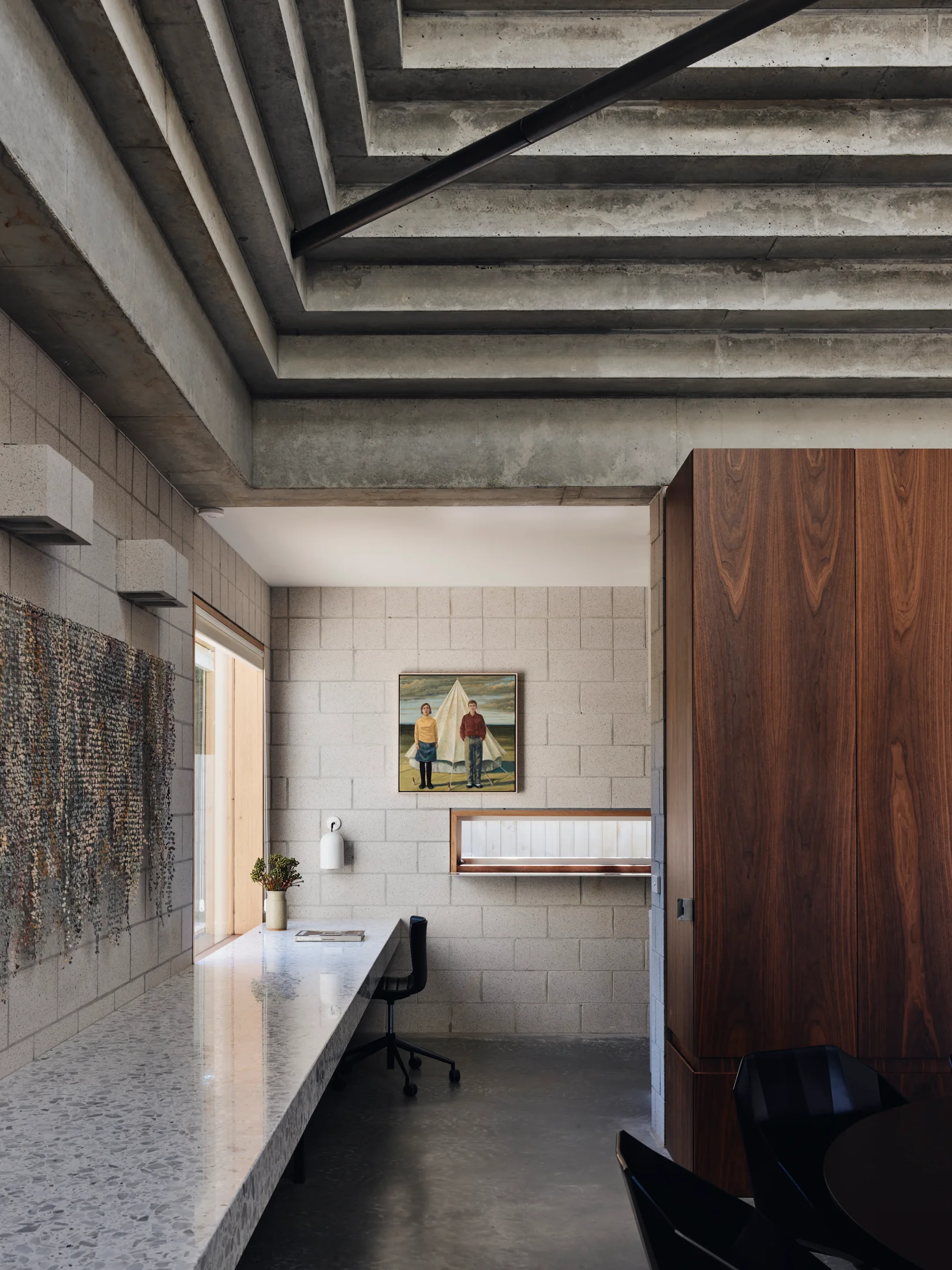 A modern home office featuring a long terrazzo-style desk, a black office chair, exposed concrete block walls, and a stepped concrete ceiling.