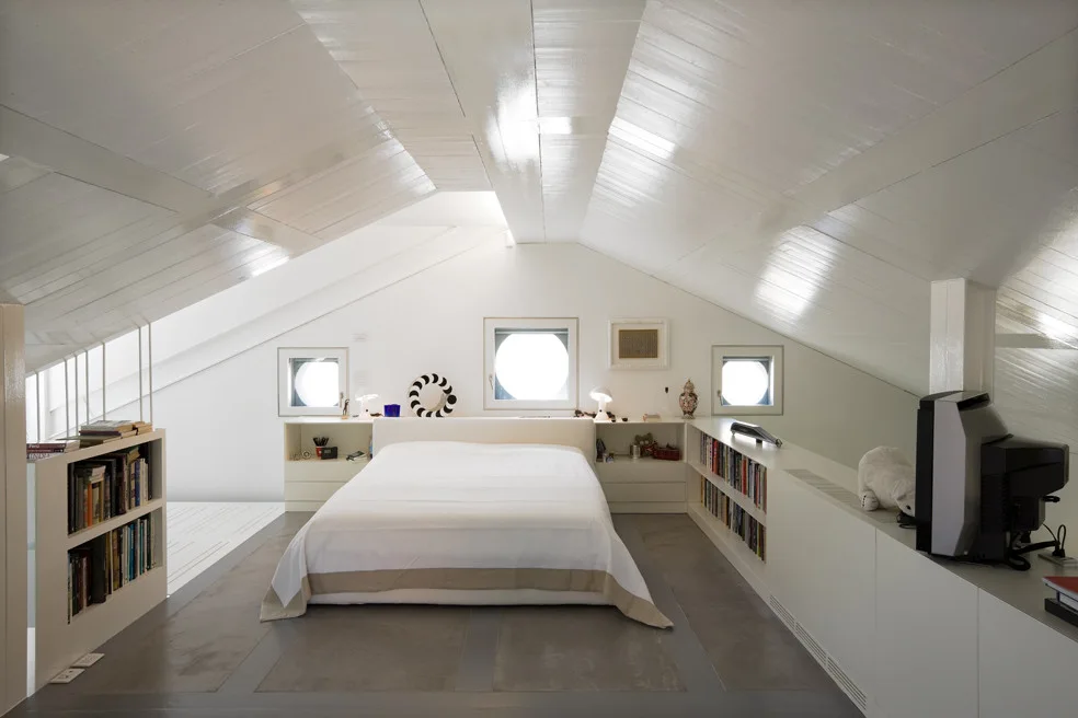 An attic bedroom with a white bed, low built-in storage wrapping the perimeter, three circular windows, and a slanted white plank ceiling.