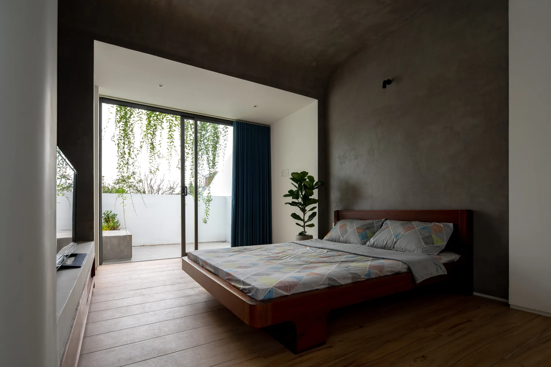 A minimalist bedroom features a wooden platform bed against a dark grey concrete wall, with sliding glass doors leading to an outdoor terrace.