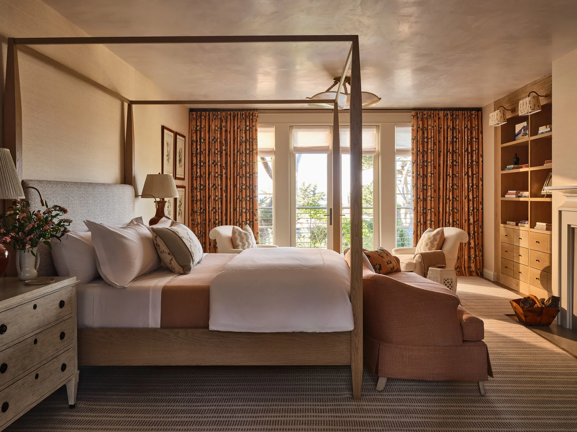 A neutral bedroom features a wooden four-poster bed, a matching upholstered bench, and patterned rust curtains by glass doors.