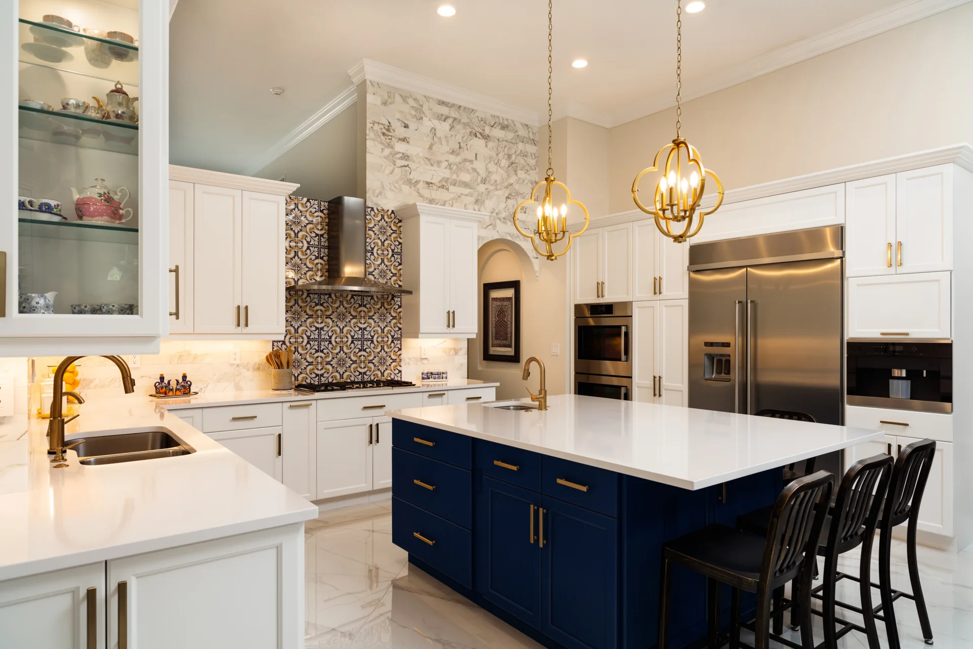 A spacious transitional kitchen features a navy blue island, white cabinets, gold pendant lights, and a patterned tile backsplash behind the stove.