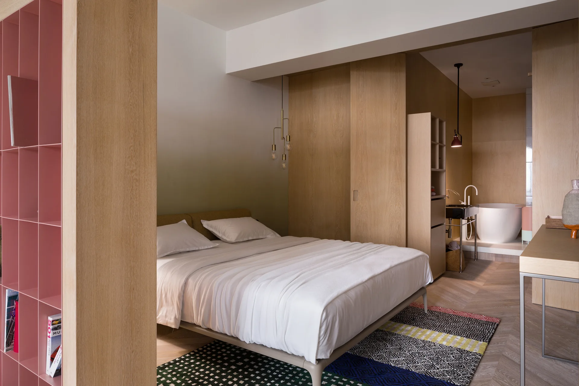 A contemporary bedroom features a bed with white bedding, a pink metal shelving unit, wood-paneled walls, and an integrated bathroom in the background.