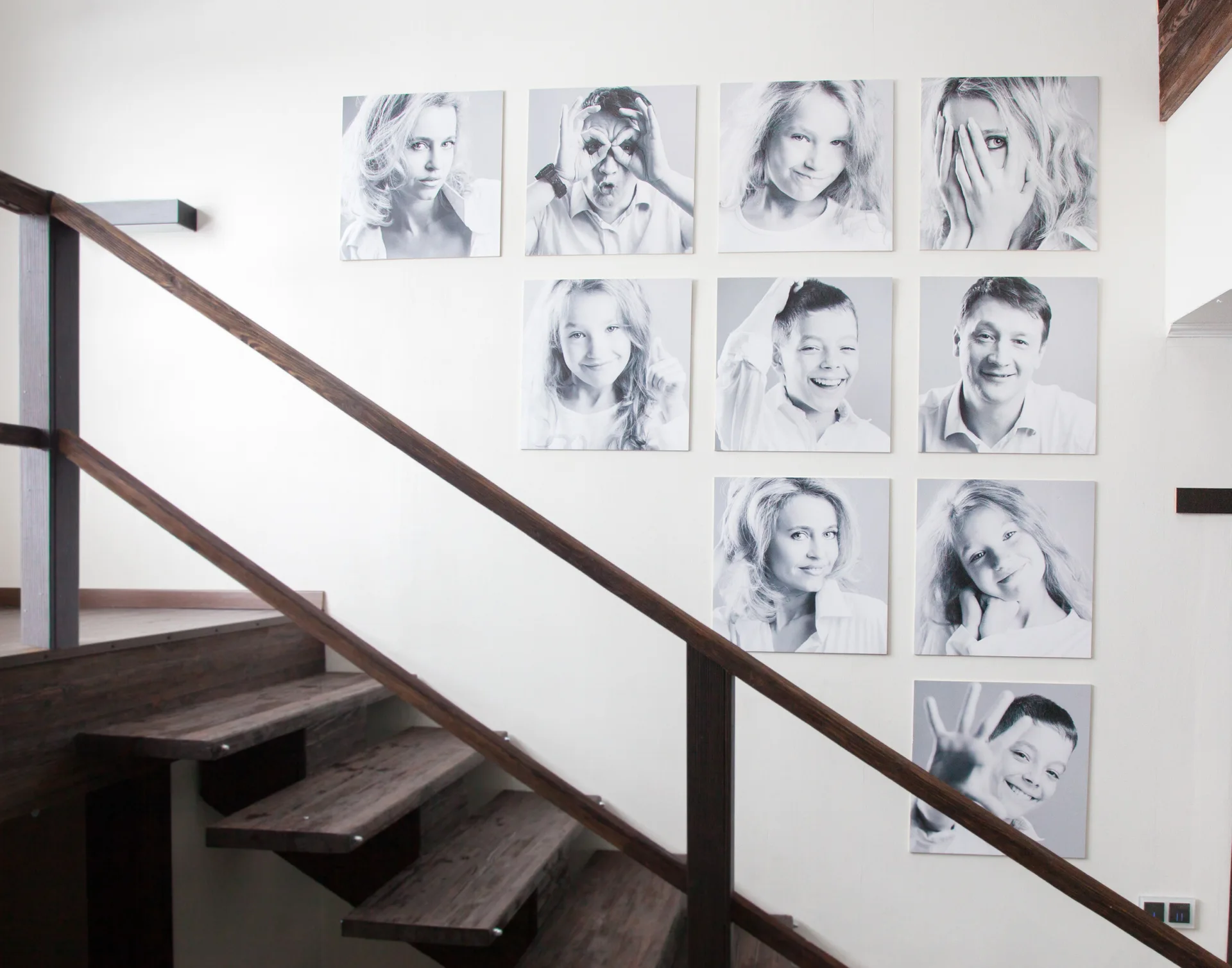 A dark wood staircase ascends next to a white wall decorated with a grid of square black and white family portraits.