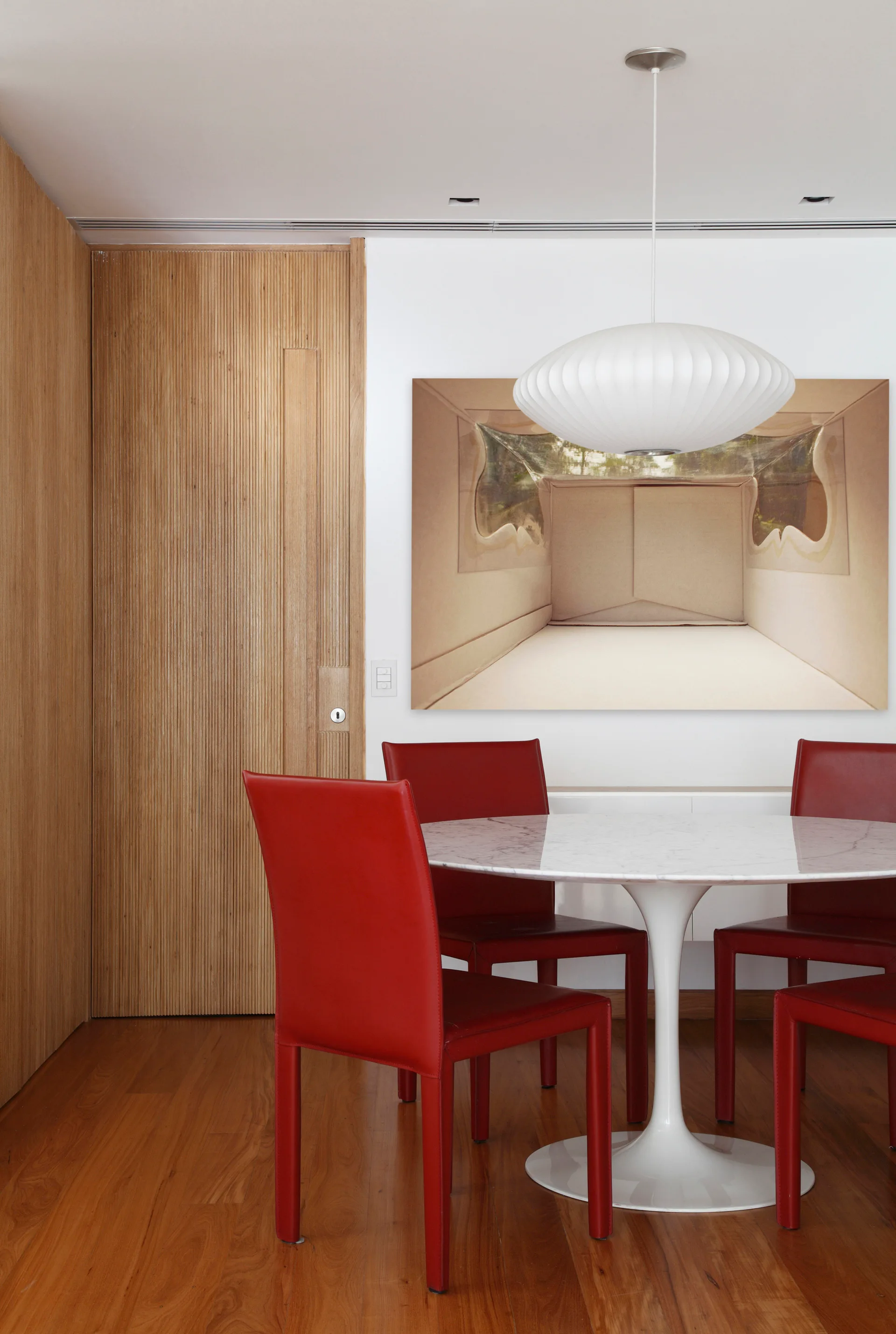 A modern dining area features a white circular pedestal table with four red chairs, a white saucer pendant light, and a fluted wood accent wall.