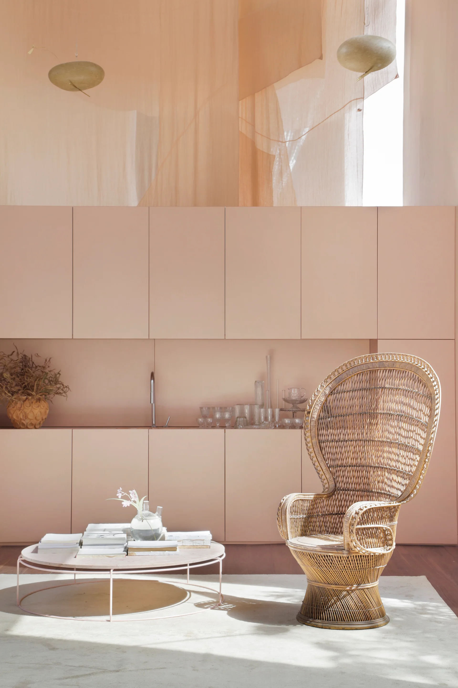 A woven rattan peacock chair sits beside a low round coffee table in front of seamless pastel pink minimalist kitchen cabinetry.