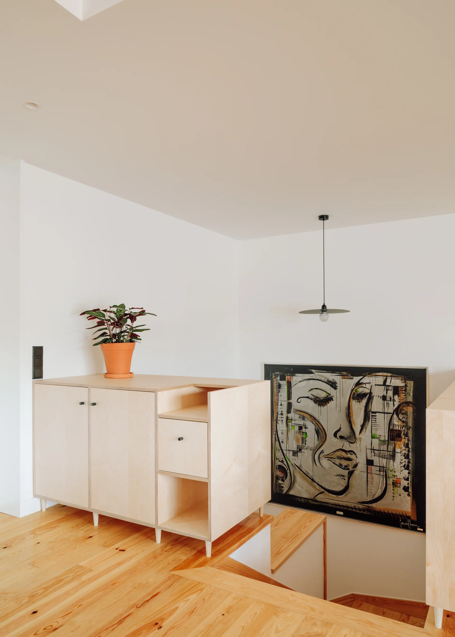 A custom light wood plywood cabinet stands on a wooden staircase landing next to a large framed painting and a minimalist black pendant light.