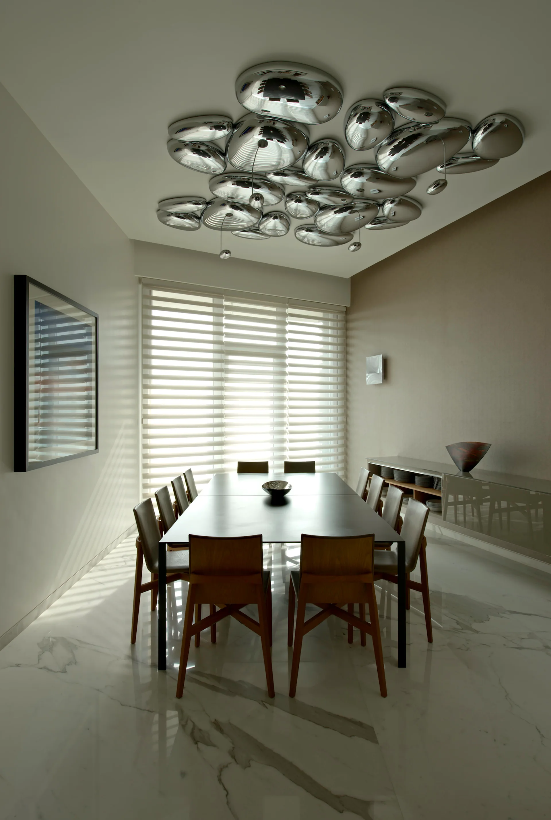 A contemporary dining room features a dark table with wooden chairs, white marble-patterned tile floors, and a clustered chrome ceiling light installation.