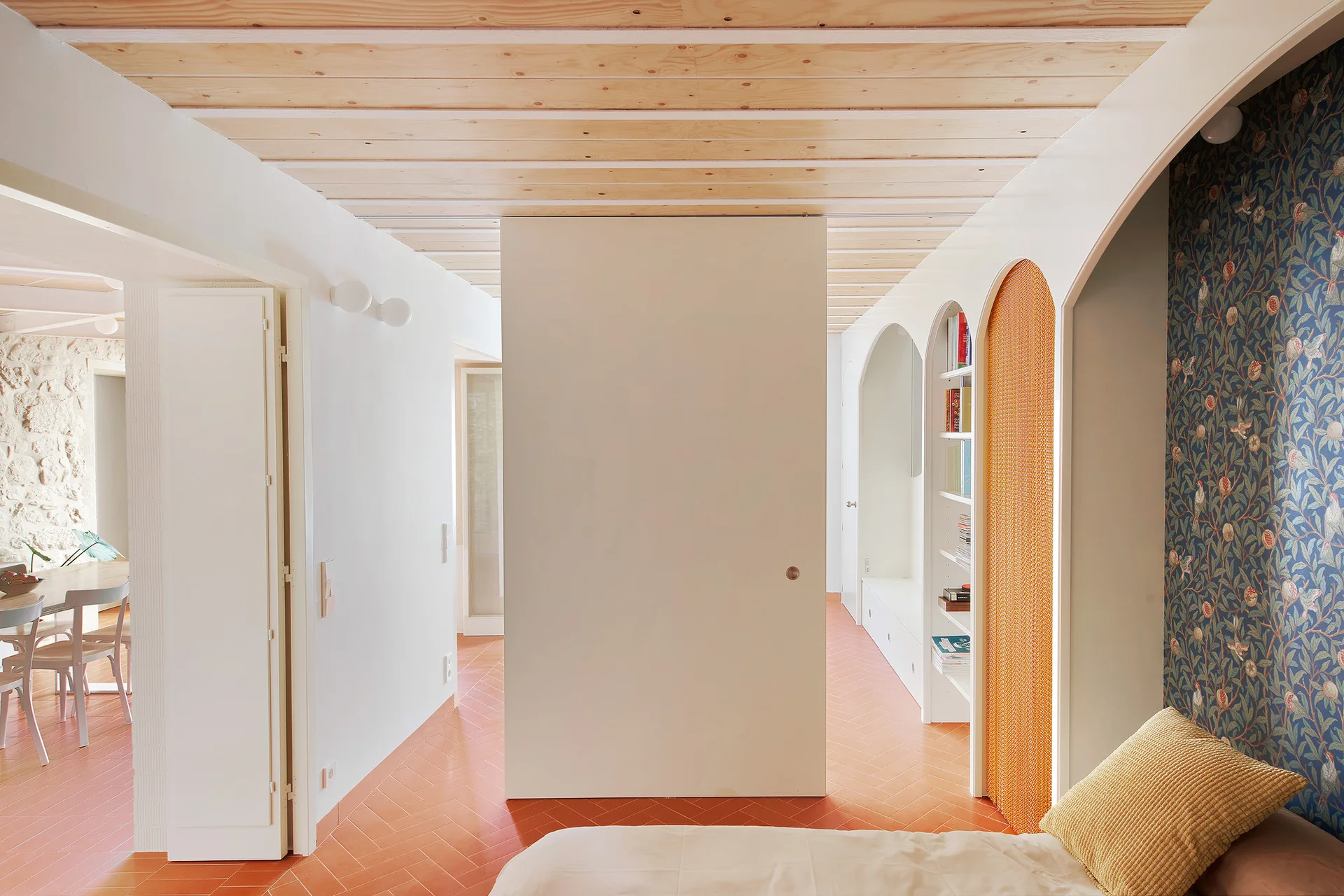 A residential hallway features terracotta herringbone floor tiles, a light wood plank ceiling, a white sliding door, and built-in arched shelving.