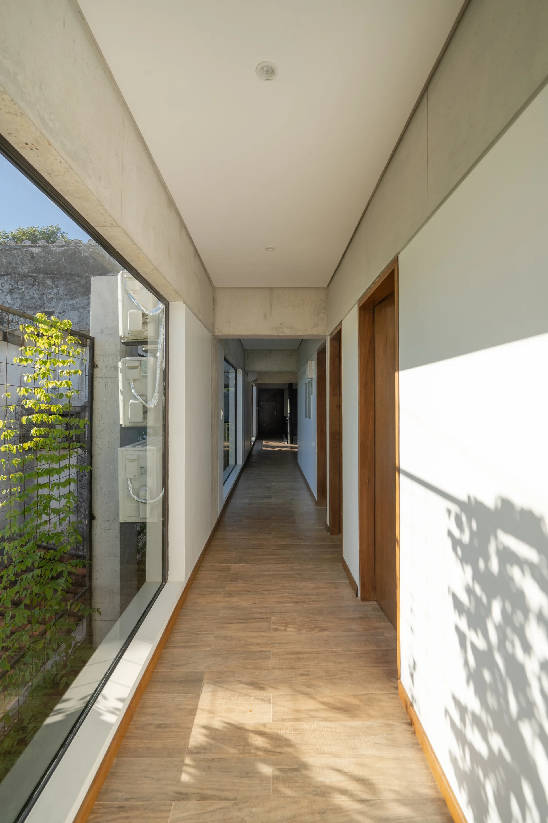 A long minimalist hallway features wooden plank flooring, matching wood doors, and a large side window beneath an exposed concrete beam.