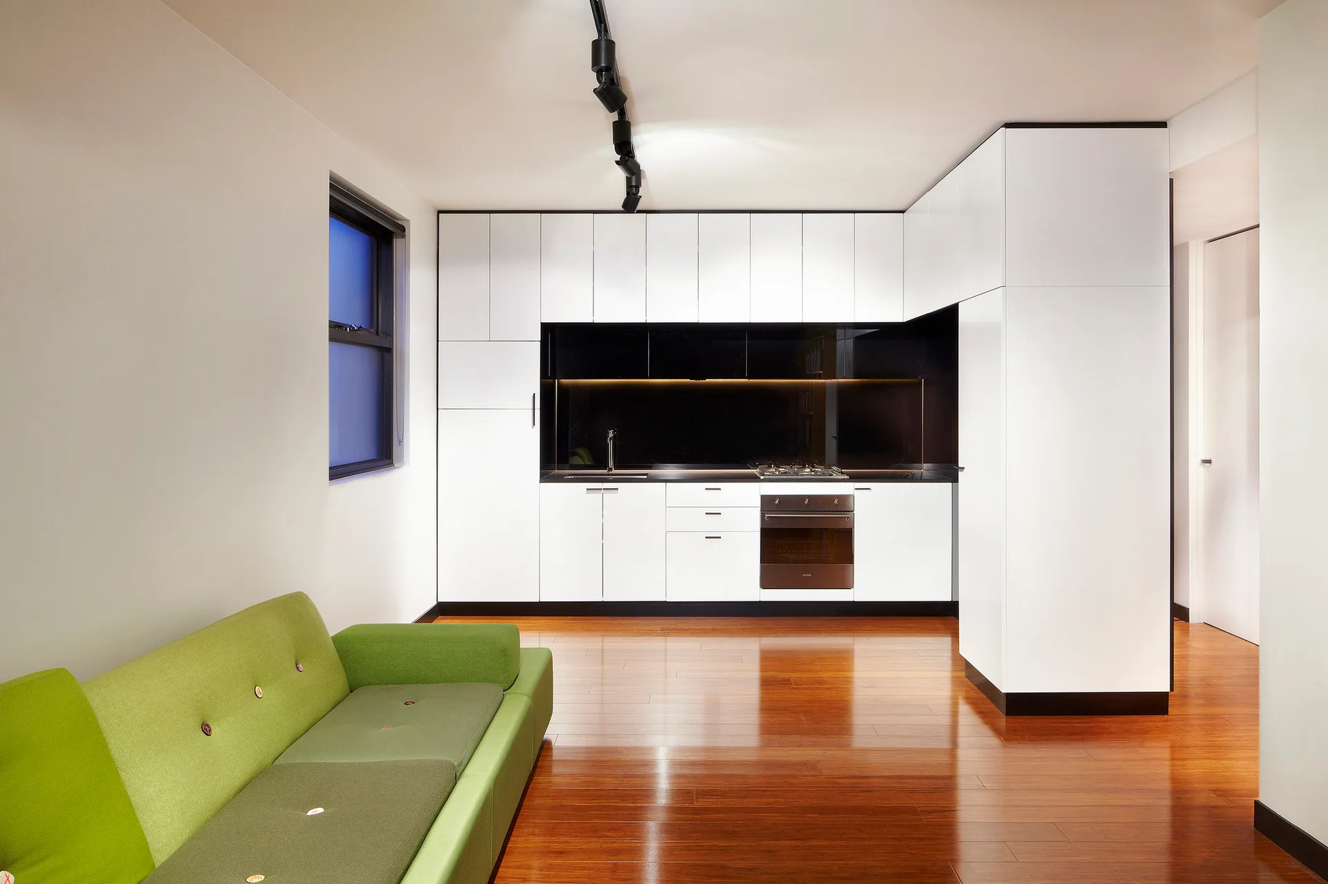 A minimalist white kitchen features a recessed black backsplash, polished wood flooring, and a green sofa in the foreground.
