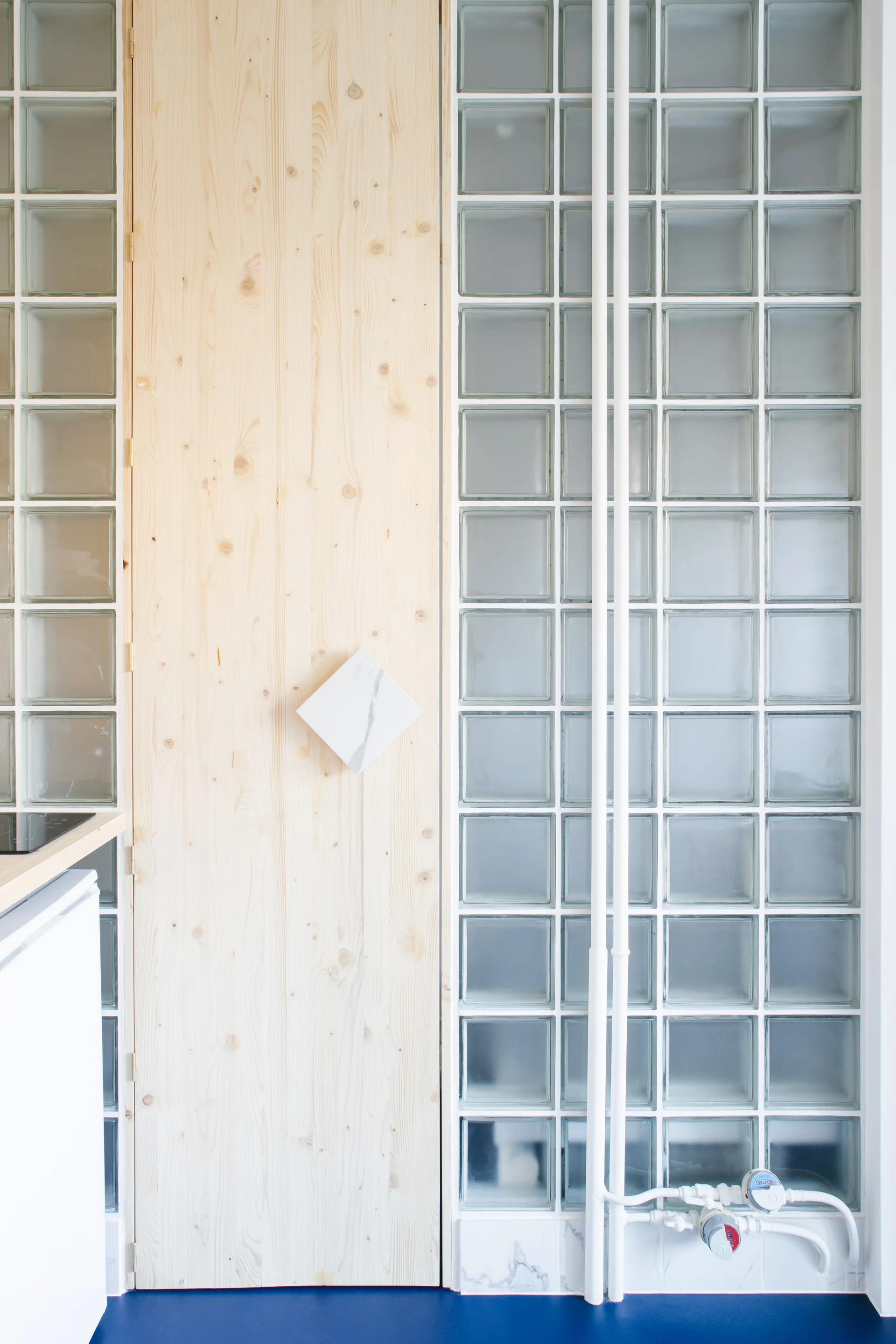 A tall pine wood panel door featuring a diamond-shaped white marble handle is set within a translucent glass block wall above a blue floor.