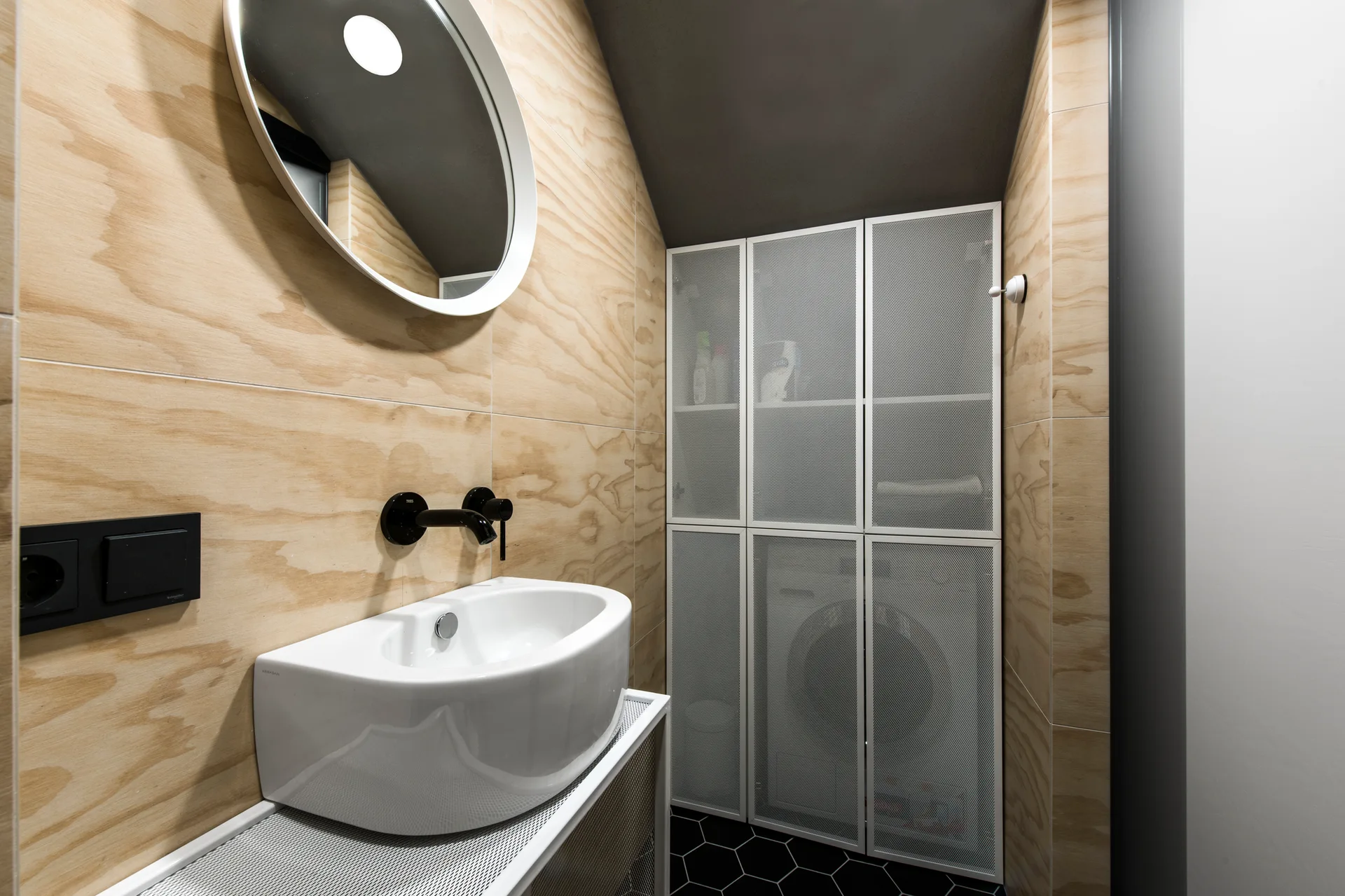 A small bathroom features light wood wall paneling, a white vessel sink with a black faucet, and a tall white metal mesh storage cabinet.
