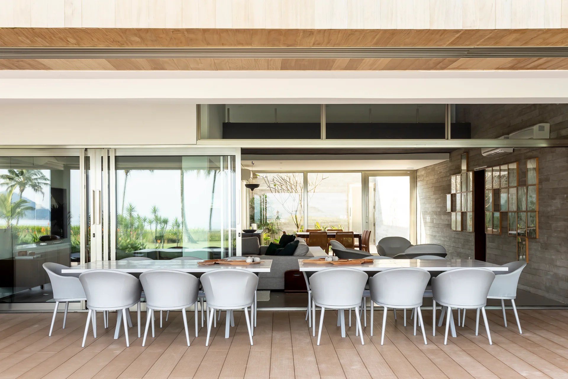 A modern outdoor patio features two long white dining tables with grey chairs, open to an indoor living area through large sliding glass doors.