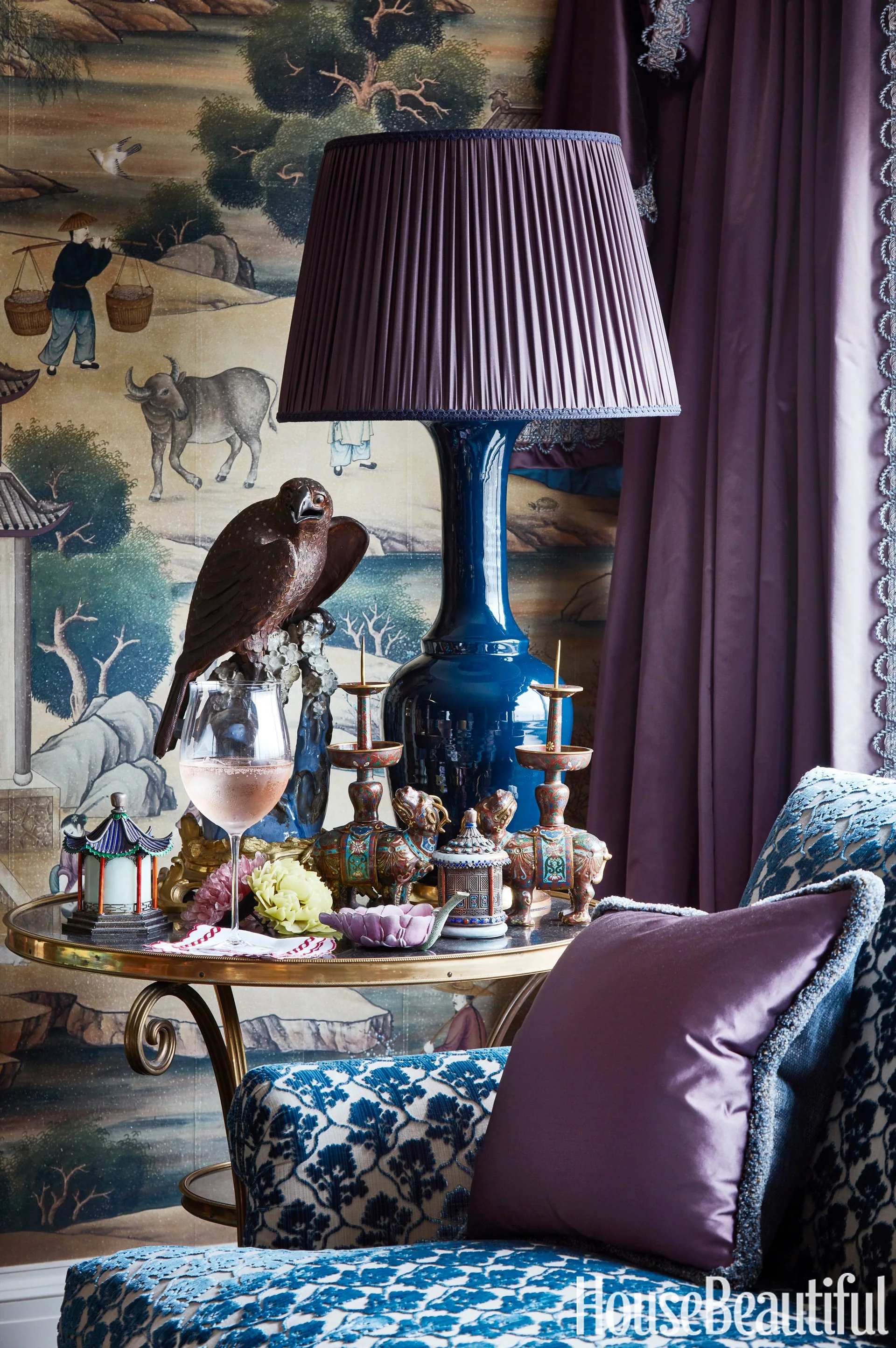 A brass side table holds a glossy blue ceramic lamp, decorative candlesticks, and a bird statue in front of chinoiserie wallpaper.