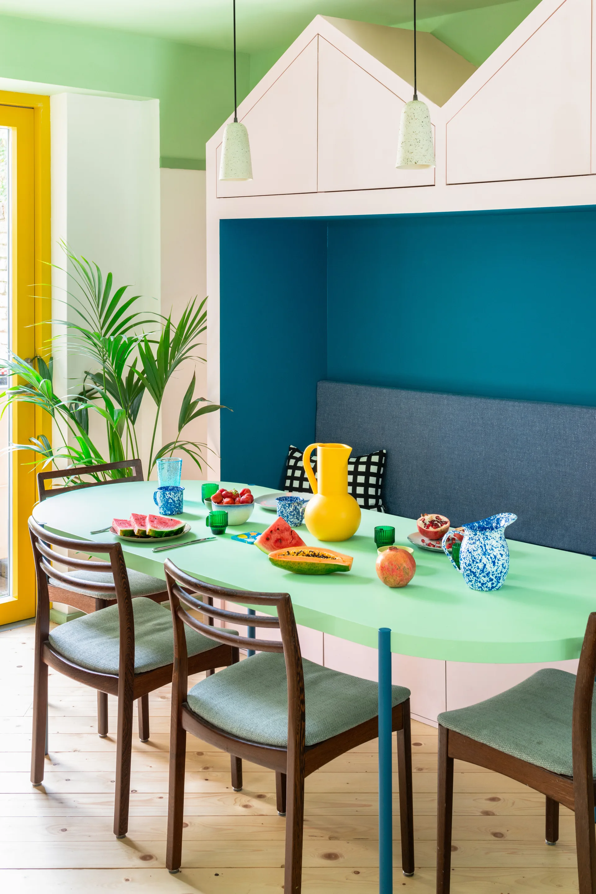 A colorful dining room features a mint green oval table, wooden chairs, a dark blue banquette niche, and pink house-shaped overhead cabinets.