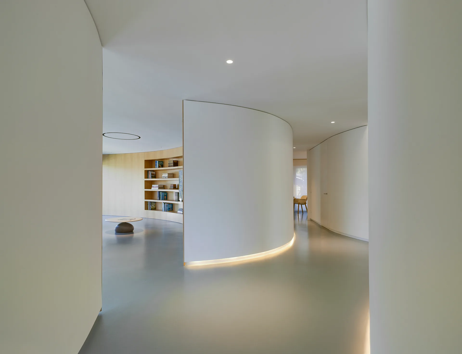 A minimalist interior features a curved white partition wall with baseboard lighting, smooth grey floors, and built-in wood shelving in the background.
