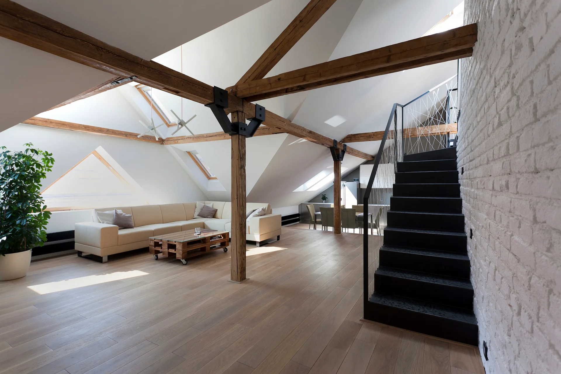 A spacious attic loft living room featuring exposed wood ceiling beams, a white brick wall, a cream sectional, and a modern black staircase.