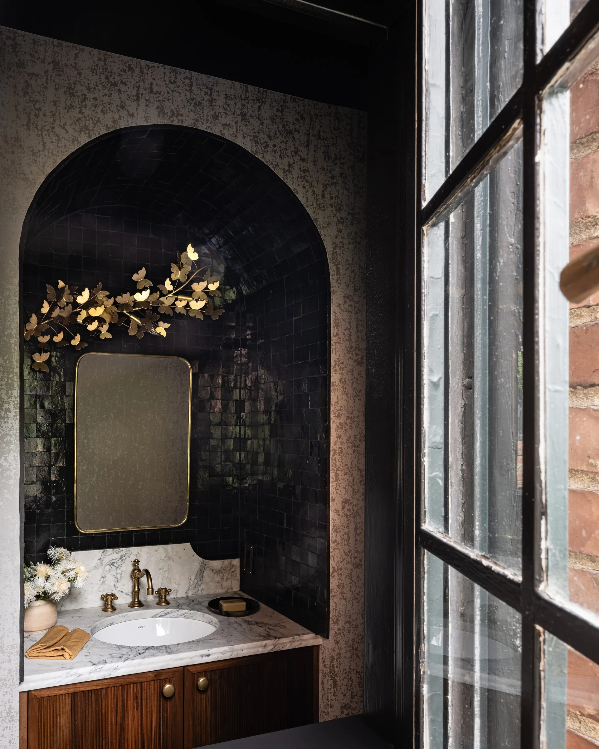 A powder room vanity with a white marble countertop, fluted wood cabinet, and an arched niche lined in glossy black tiles.