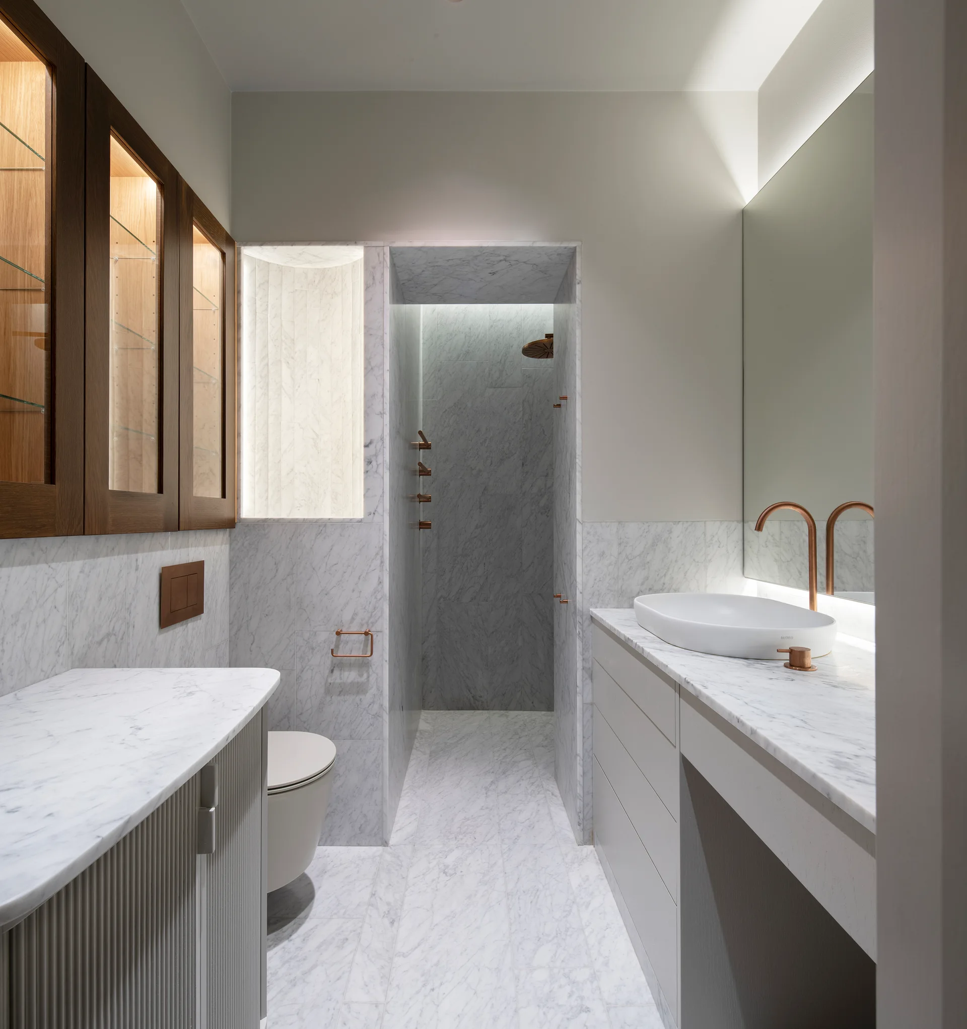 A minimalist bathroom with white veined marble walls and floors, featuring a white vessel sink on a vanity, copper faucets, and dark wood upper cabinets.