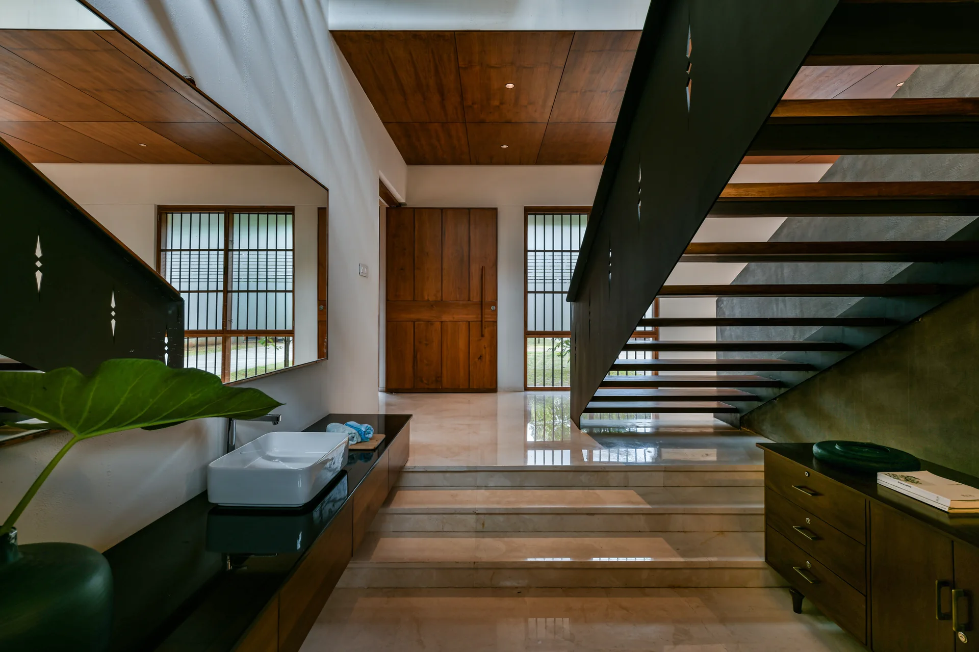 A modern entryway features a black metal staircase with open wood treads, a polished stone floor, wood ceiling panels, and a wall-mounted vanity sink.