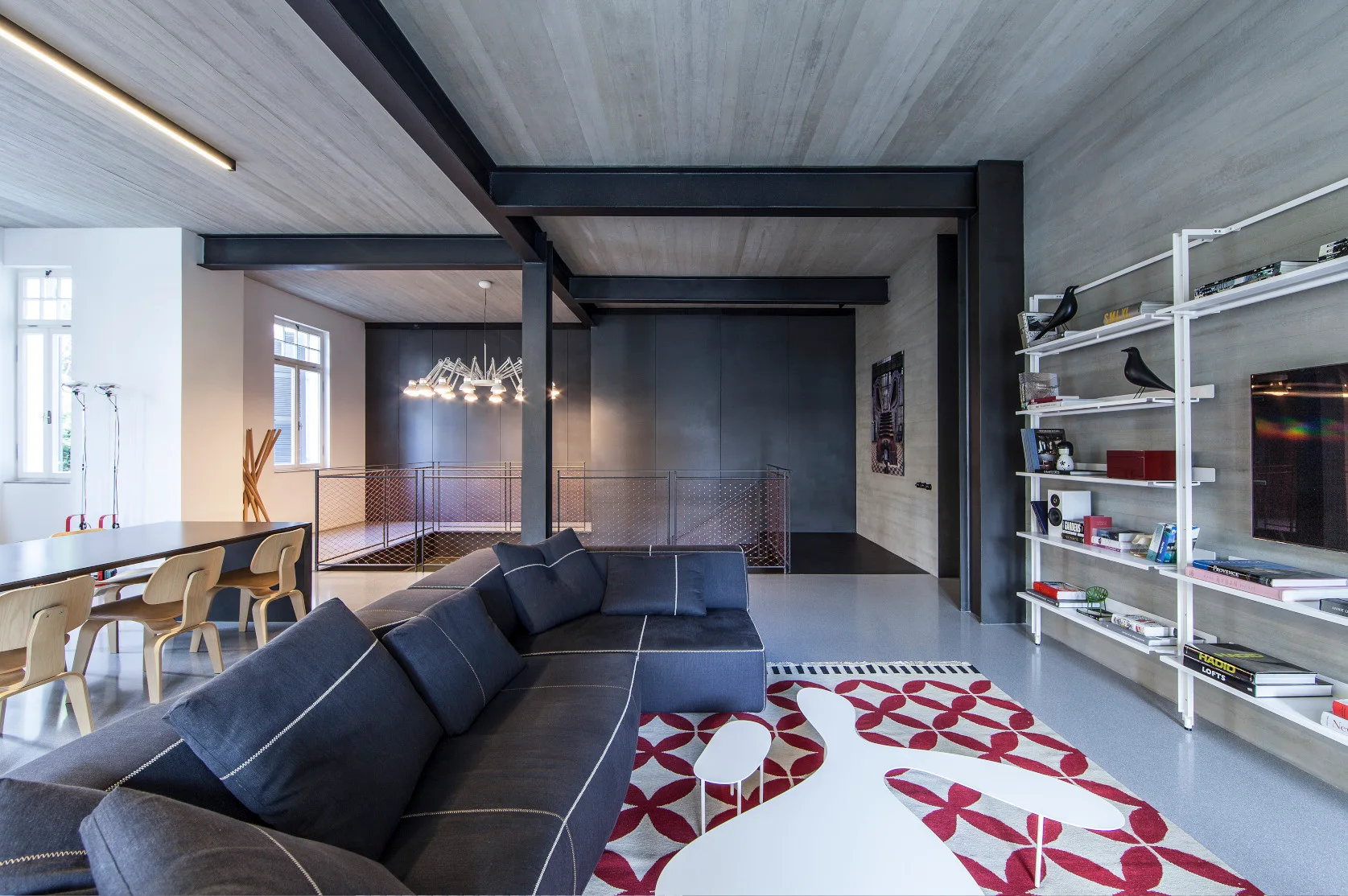 A modern industrial living room features a dark gray sectional sofa, a bold red geometric patterned rug, and exposed black steel ceiling beams.