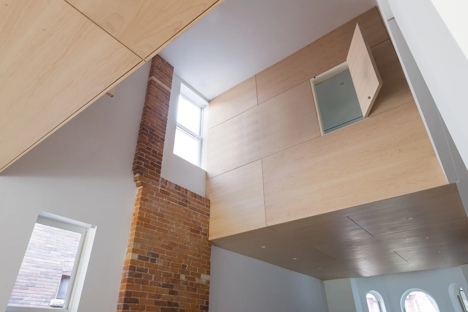 A high-ceilinged room features light wood paneling on the upper wall and ceiling, an exposed red brick column, and white walls with windows.