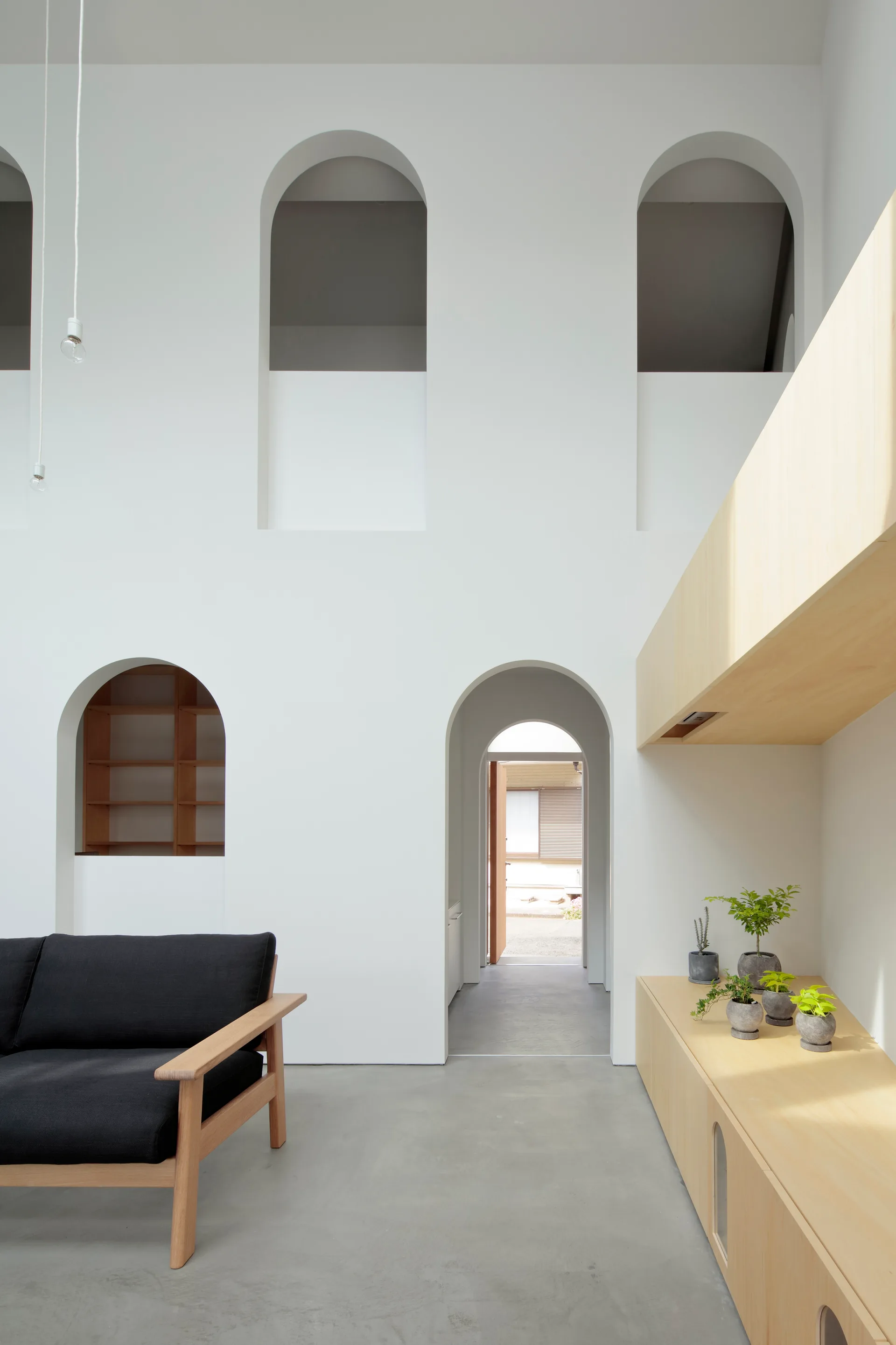 A minimalist living room with a concrete floor, a black sofa with a wood frame, a light wood console table, and several arched wall openings.