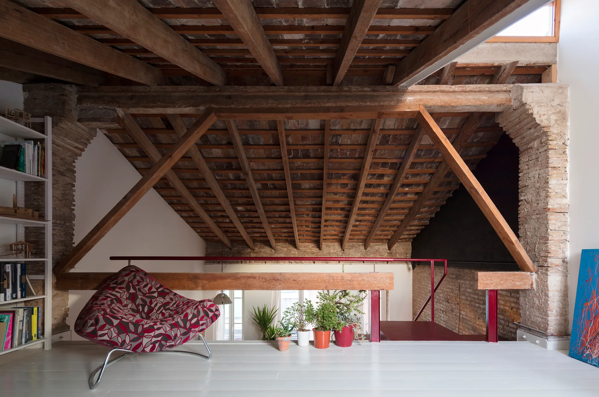 An elevated attic loft features exposed wooden rafters, a brick archway, white floors, a red-patterned armchair, and potted plants.