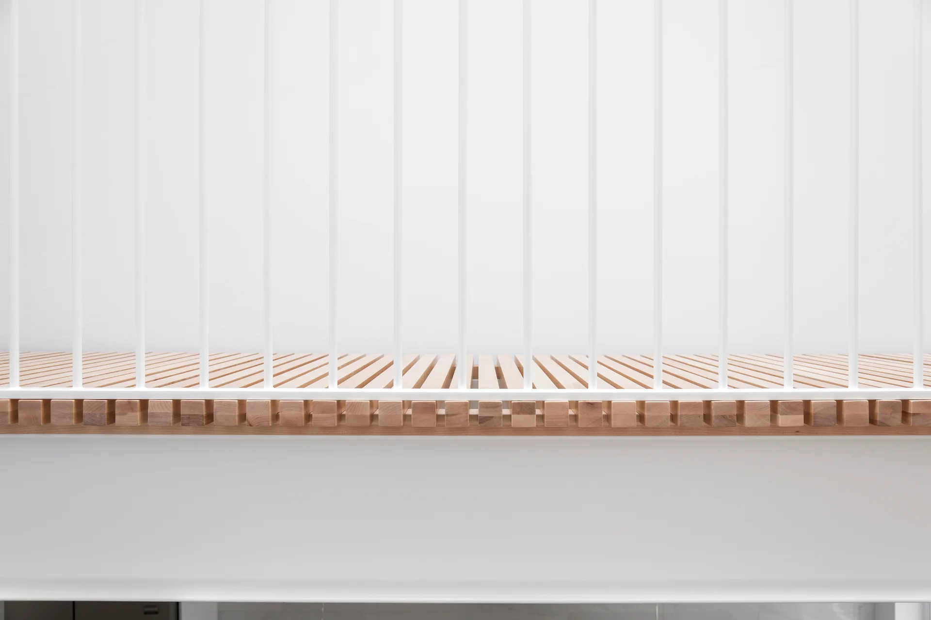 A minimalist white metal baluster railing is installed over a light natural wood slatted surface against a plain white wall.