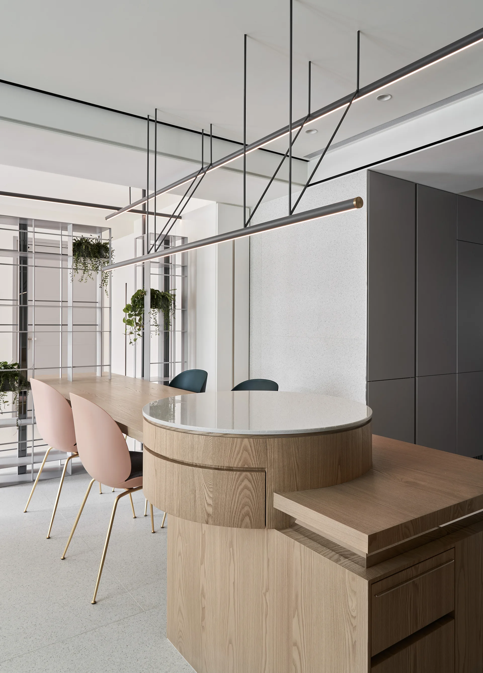 A modern kitchen featuring a custom wood island with a raised circular stone countertop, light pink dining chairs, and linear black pendant lighting.