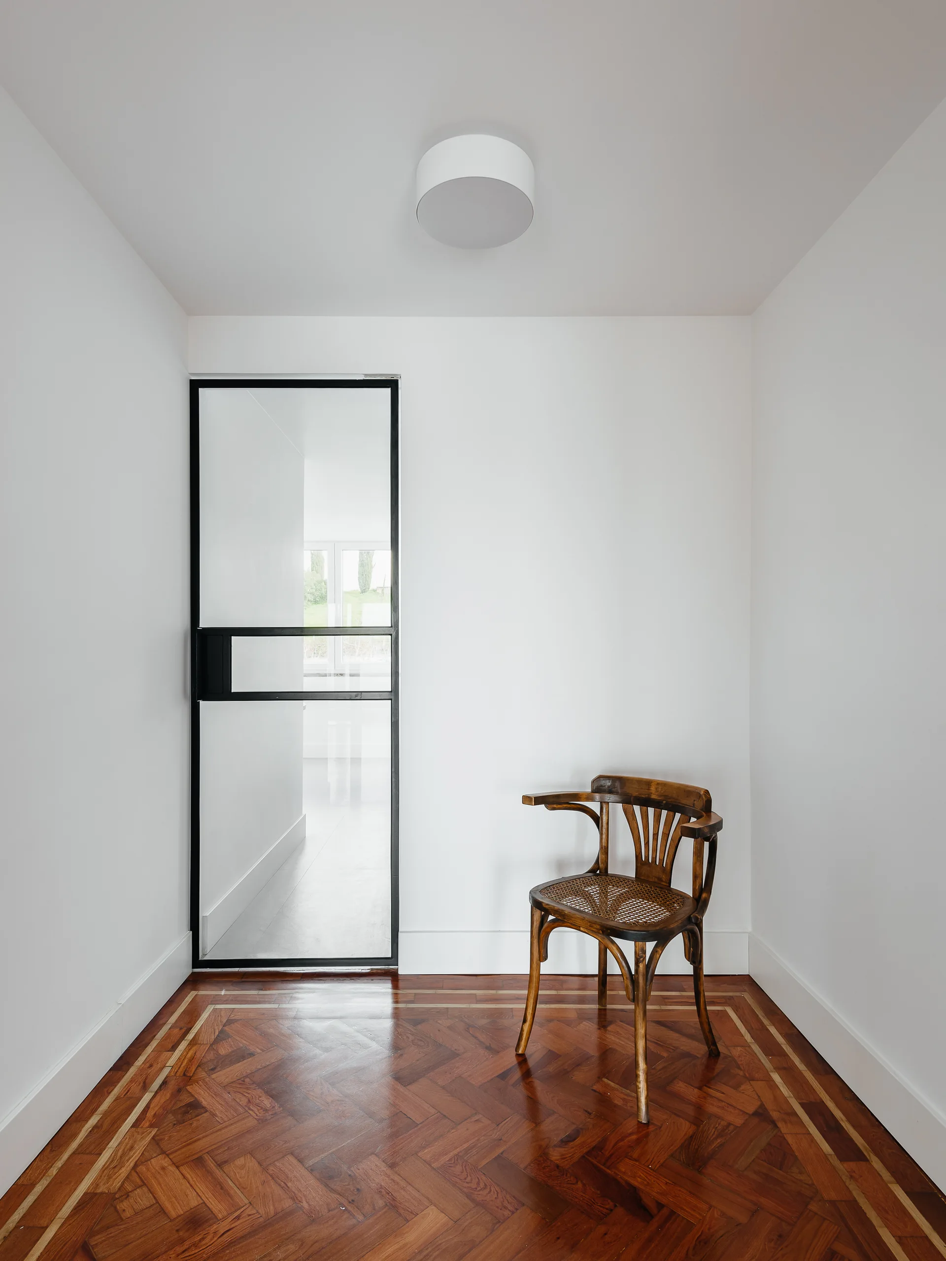 A vintage bentwood armchair sits on a rich herringbone wood floor in a minimalist white entryway with a black-framed glass door.