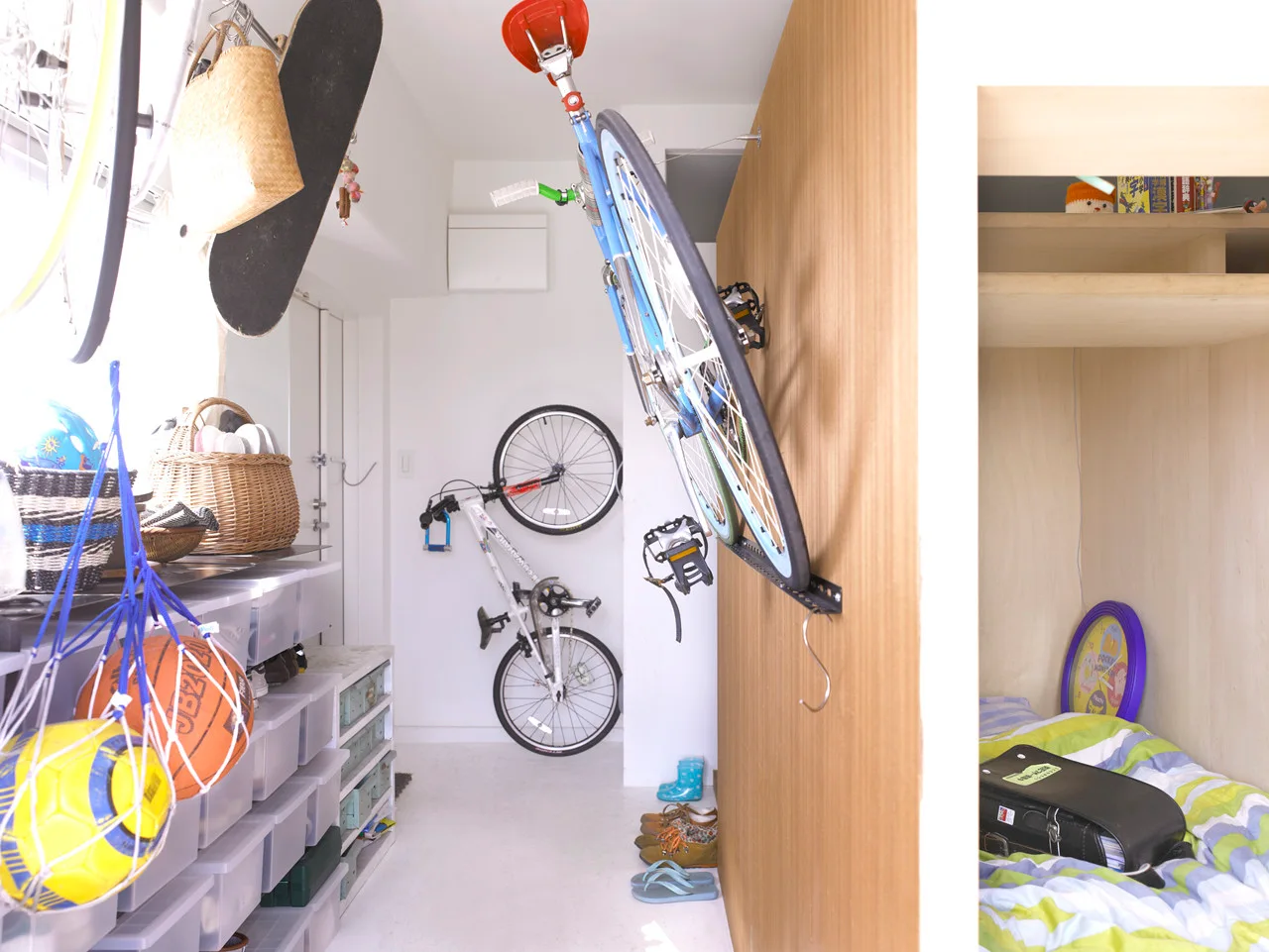A narrow white entryway features wall-mounted bicycles, a stack of plastic storage bins, and a recessed wooden sleeping nook.