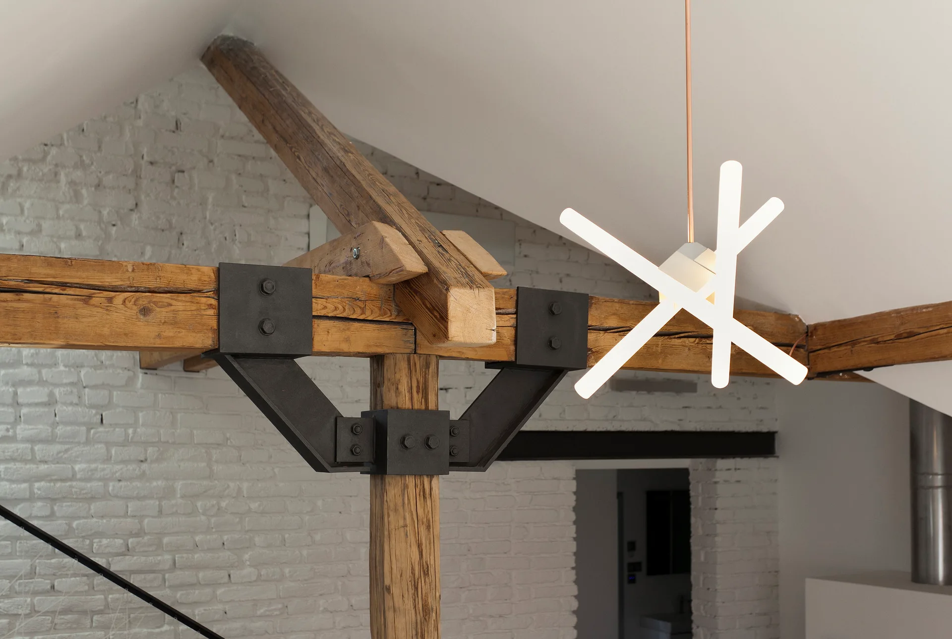 Rustic timber beams joined by black industrial steel brackets sit near a modern geometric pendant light against a white painted brick wall.