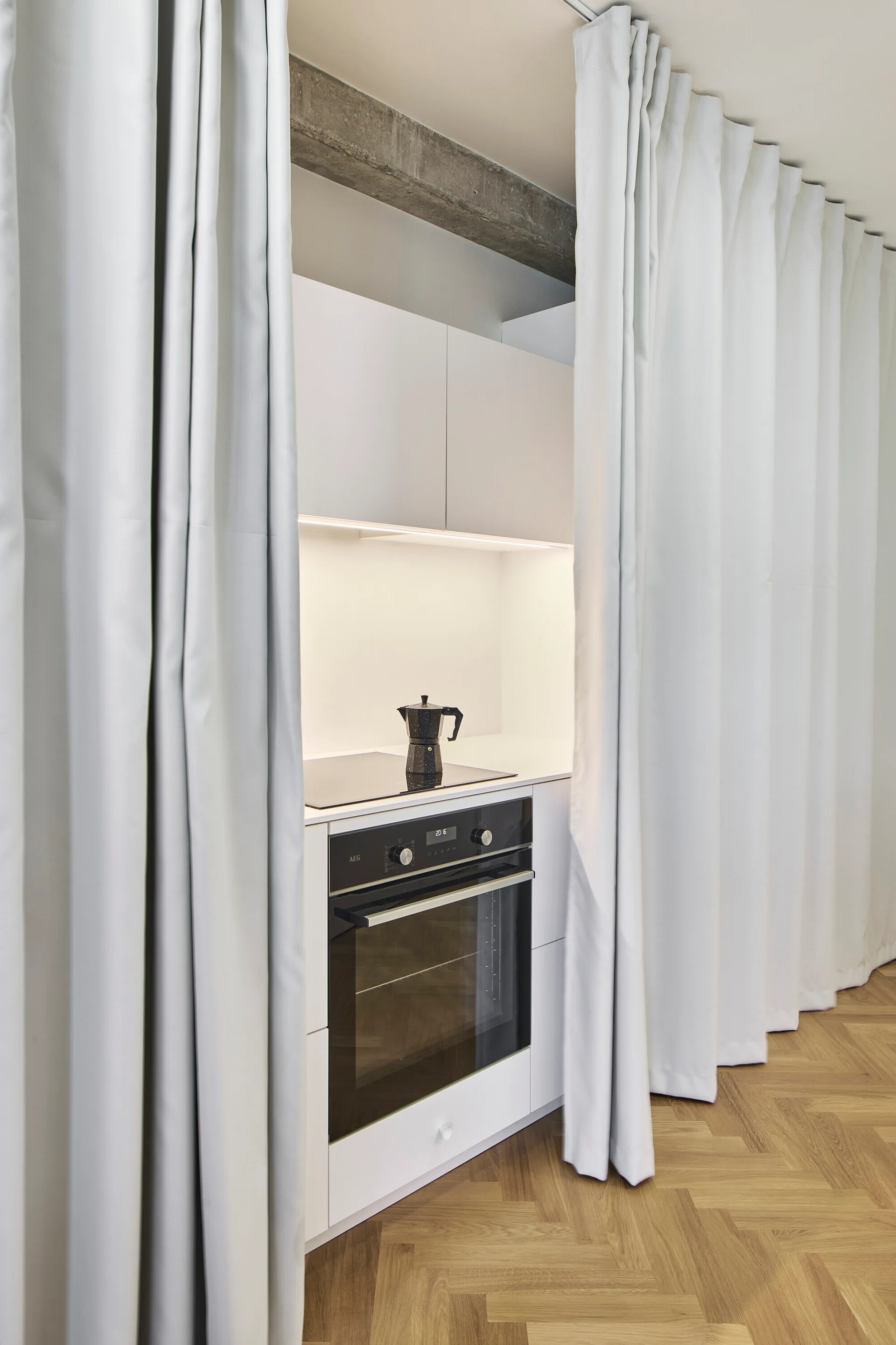 A compact white kitchenette with a black oven and cooktop is framed by long light gray curtains on herringbone wood flooring.