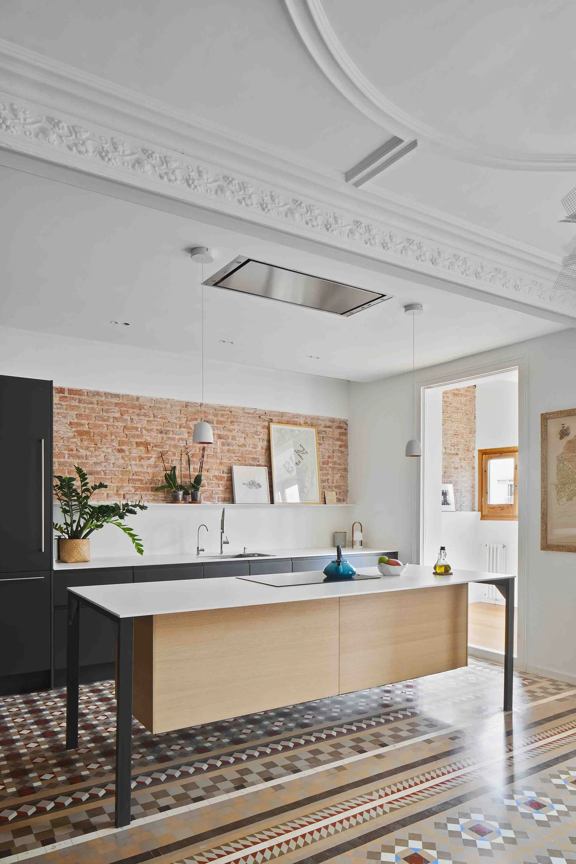 A modern kitchen featuring an island with a light wood base, dark gray cabinets against an exposed brick wall, and a patterned mosaic tile floor.
