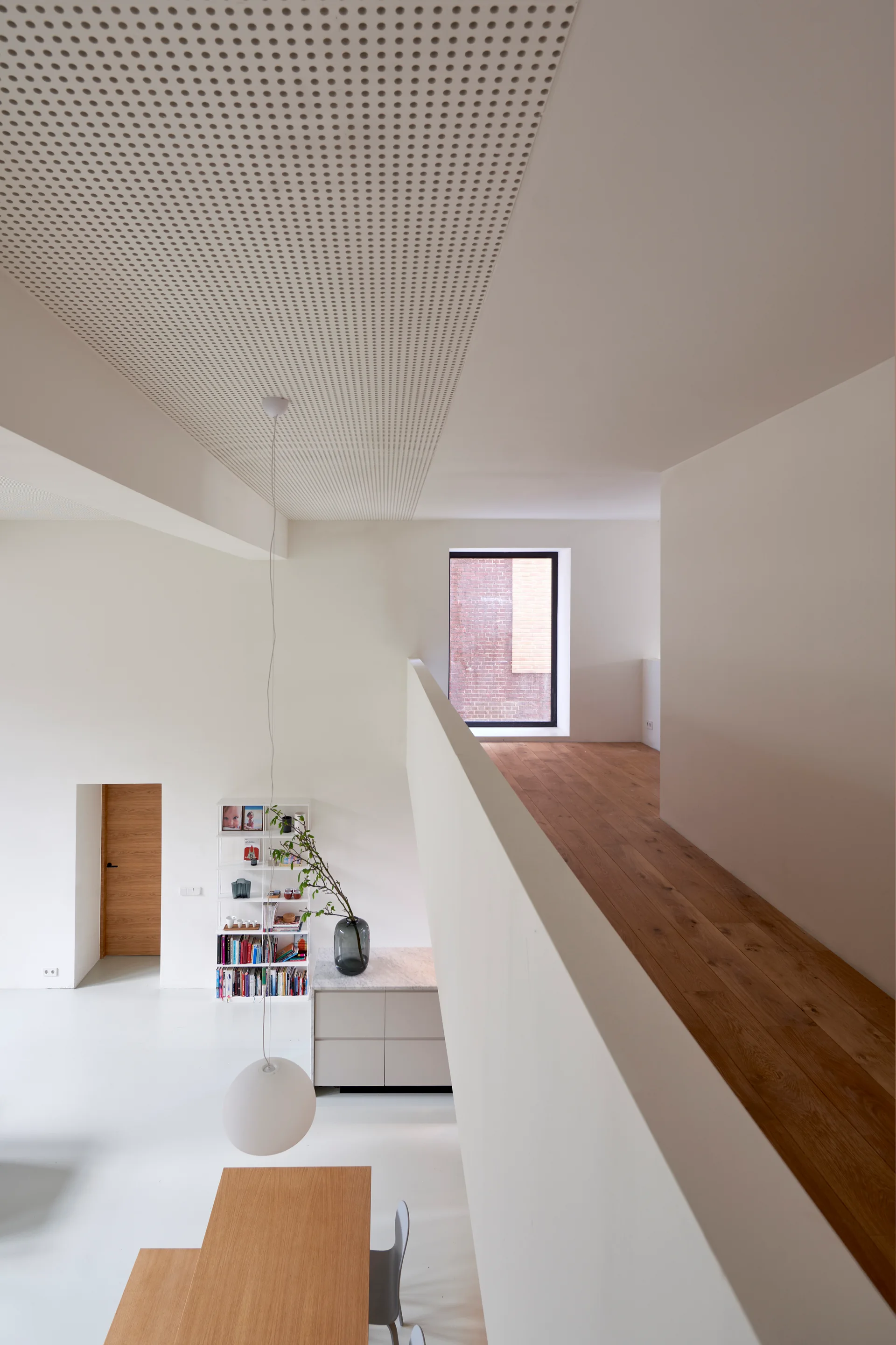 A bright minimalist loft featuring a white dining area beneath a wooden mezzanine walkway and a perforated acoustic ceiling.
