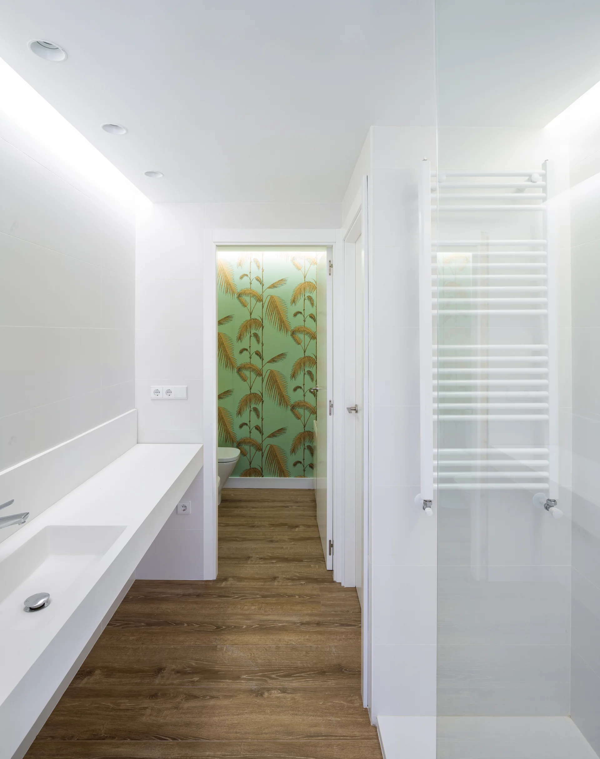 A modern white bathroom features a long integrated vanity, wood flooring, and a view into a separate toilet closet with green palm leaf wallpaper.