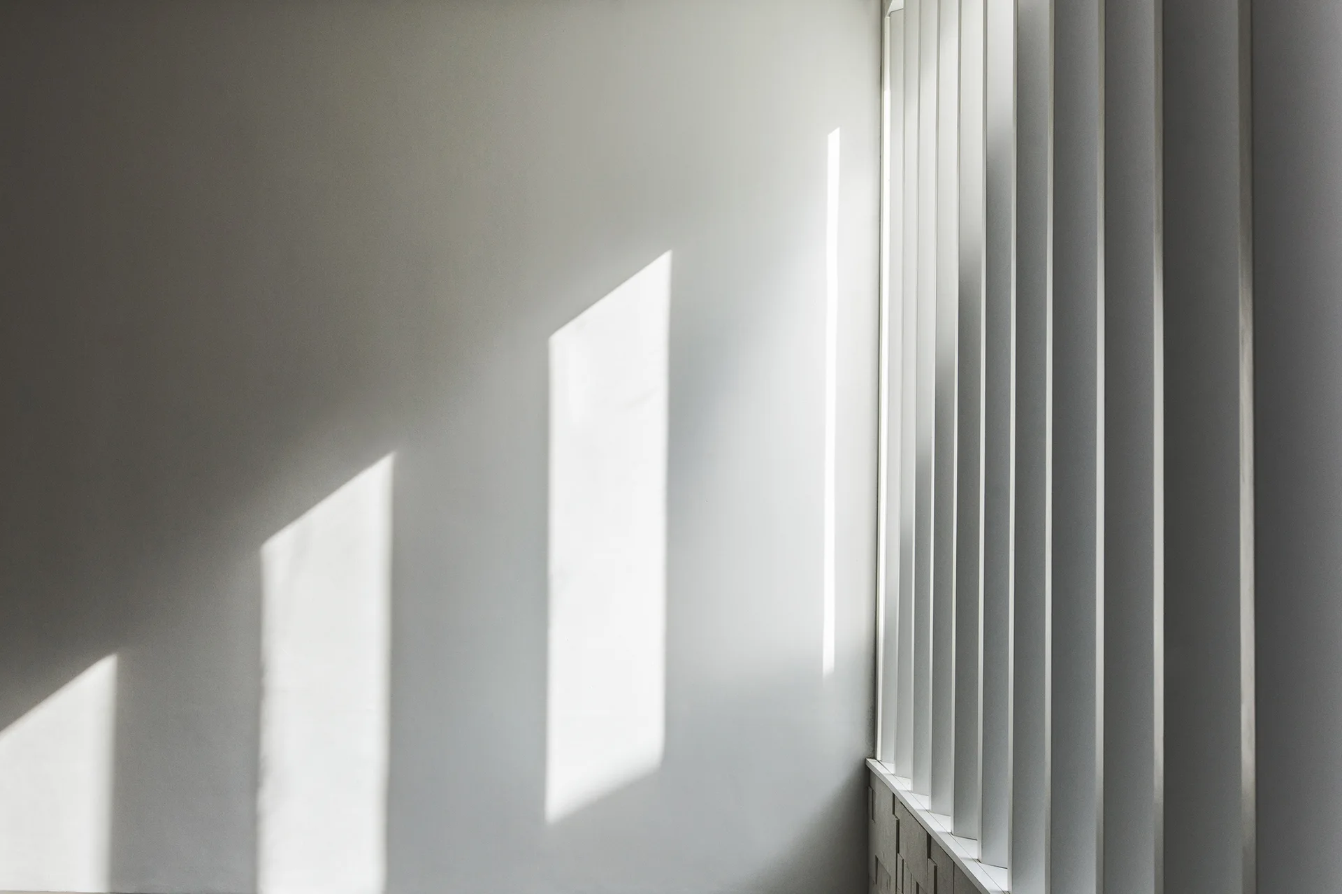 Crisp geometric shadows and light patterns fall across a smooth white wall next to a row of vertical architectural louvers.