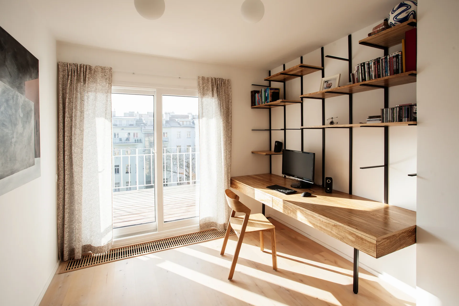 A minimalist home office features a long wall-mounted wooden desk, an open metal and wood shelving unit, and a sliding glass door opening to a balcony.