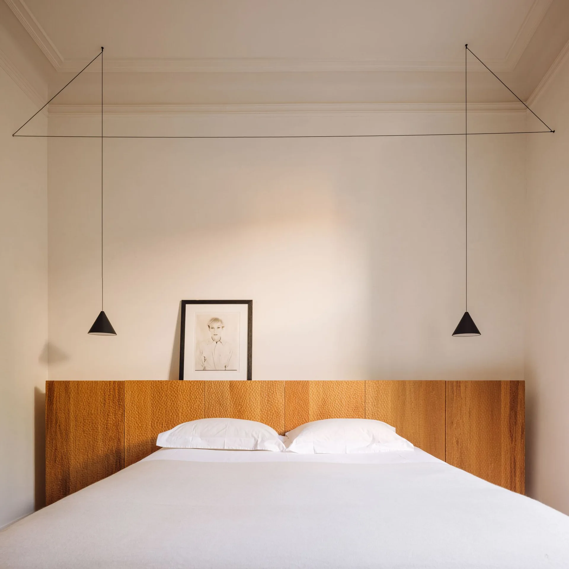 A minimalist bed with white sheets sits against a wall-to-wall carved wood headboard with two hanging black cone pendant lights.